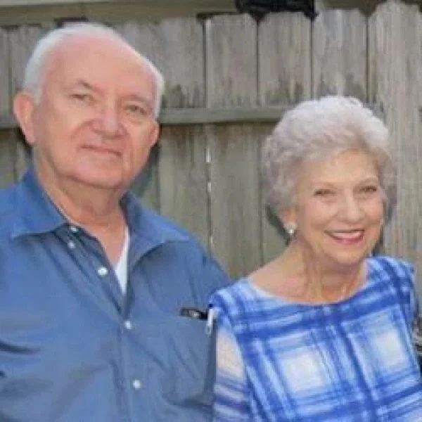 An elderly man and woman standing outside in front of a wooden fence, smiling at the camera. The man is wearing a blue shirt and the woman is dressed in a blue plaid blouse.
