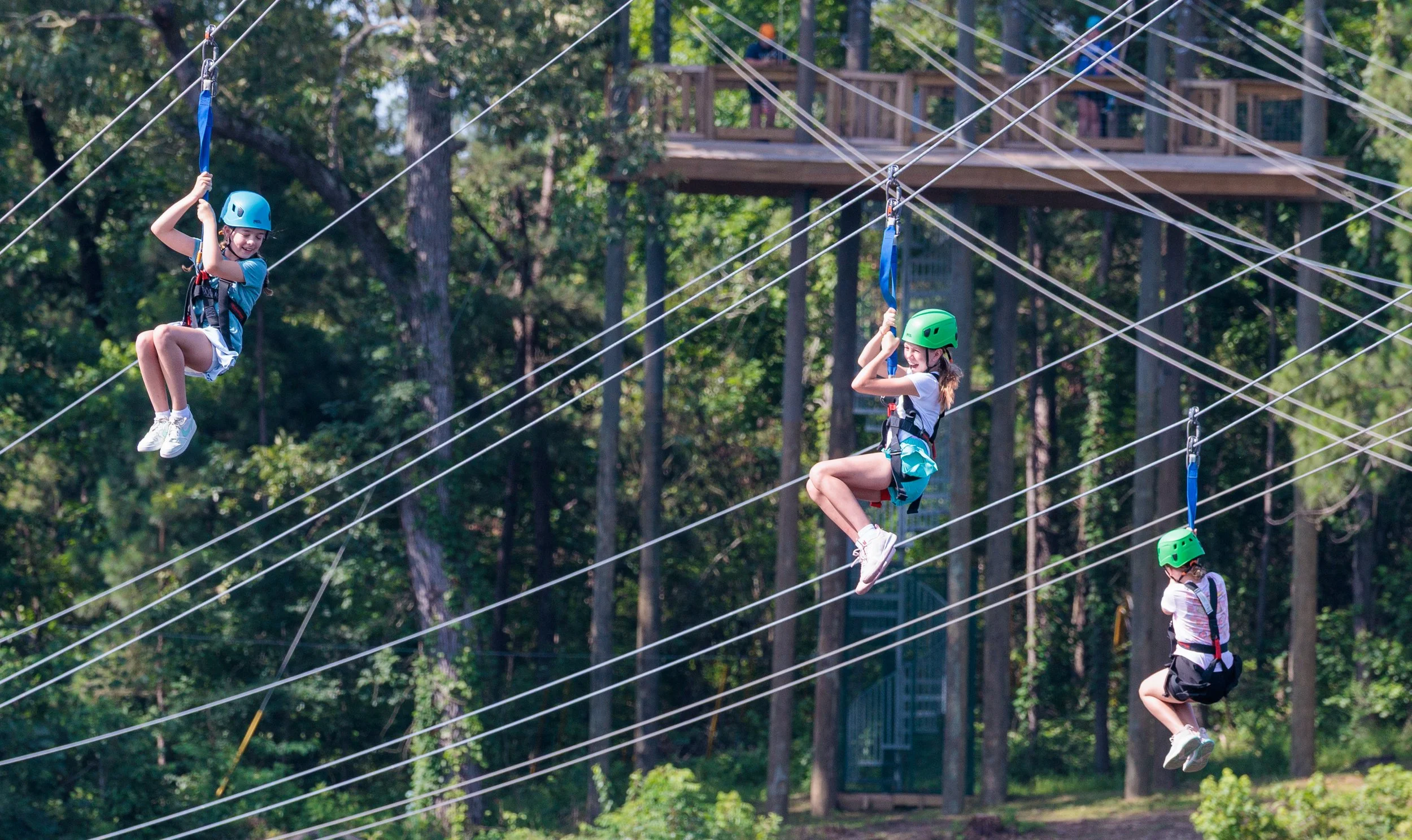 Three children wearing helmets and harnesses riding a zip line in a forest.