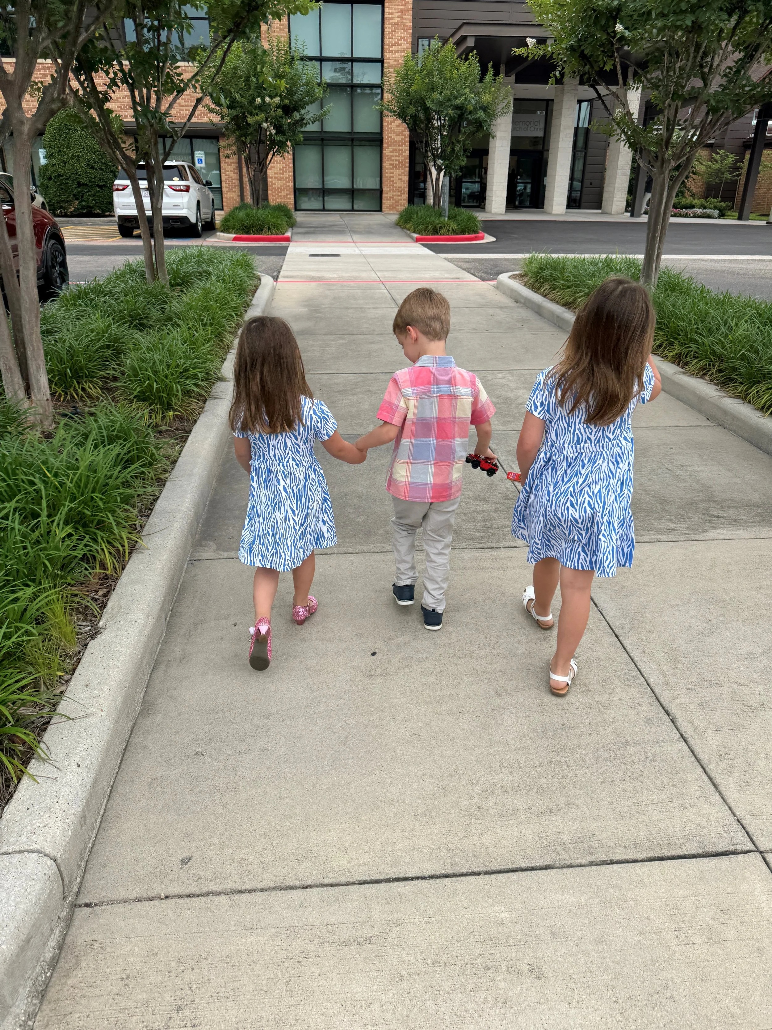 Three children walking hand-in-hand down a sidewalk in an urban area, two girls in matching blue and white dresses and a boy in a pink plaid shirt holding a toy truck.