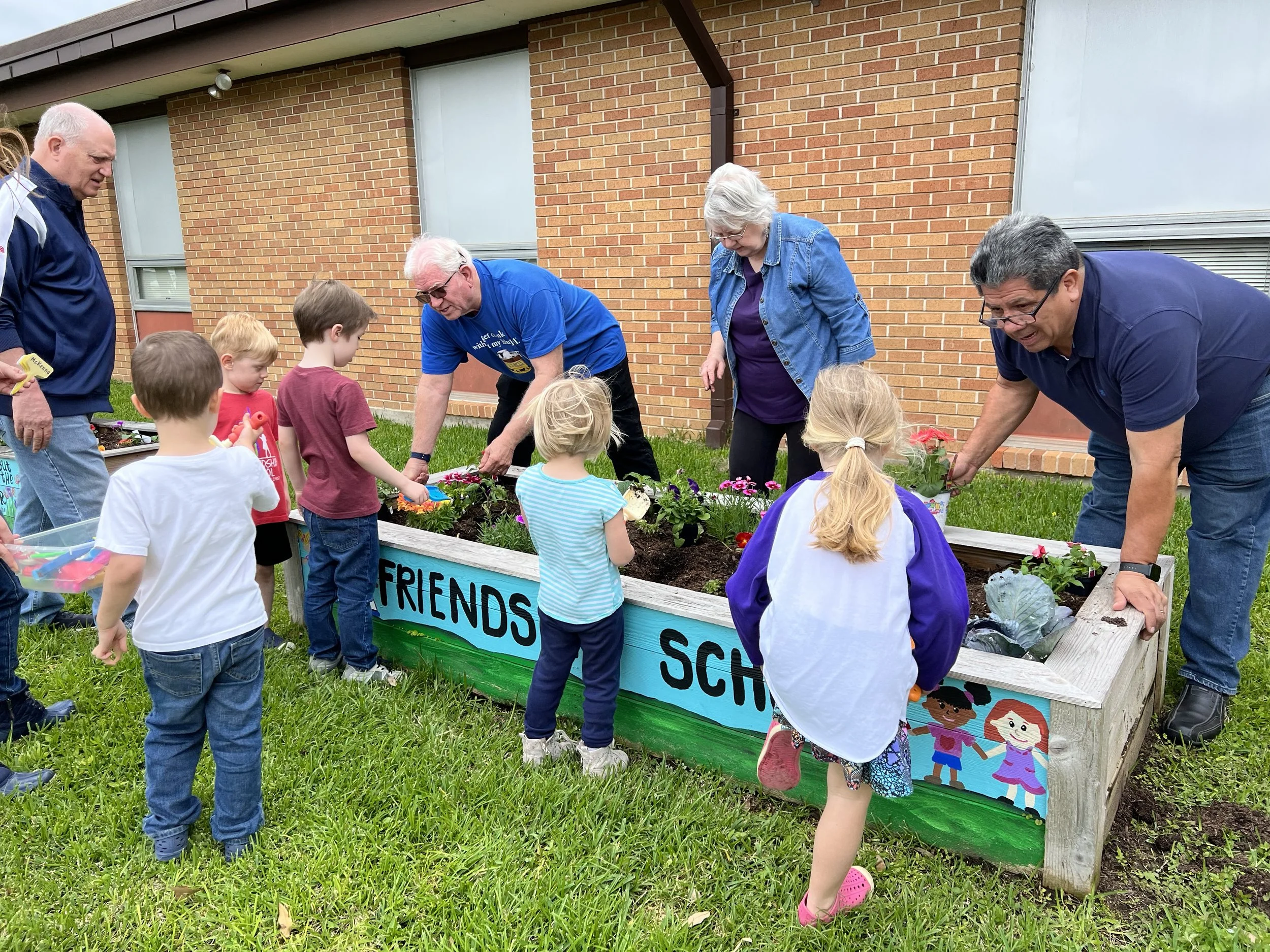 Children and adults gardening together in a raised flower bed outside a brick building.
