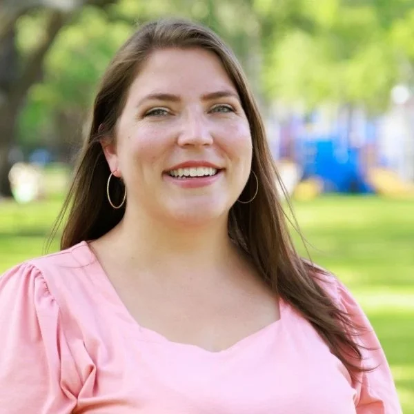 A smiling woman with long brown hair wearing a pink top and hoop earrings outdoors in a park with green trees and blurred playground equipment in the background.