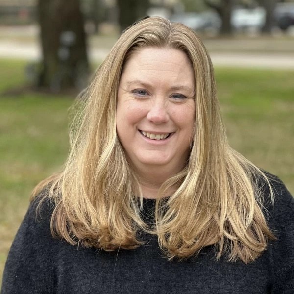A woman with long blonde hair smiling outdoors in a park or backyard.