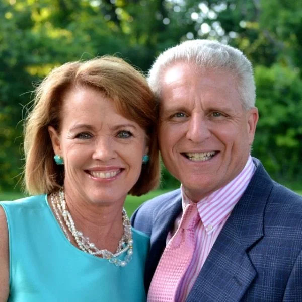 A smiling middle-aged woman with red hair wearing a turquoise dress and pearl jewelry, and a smiling middle-aged man with gray hair wearing a dark blue suit, pink tie, and pink striped shirt, standing outdoors with green trees in the background.