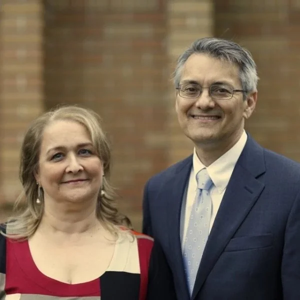 A man and woman standing side by side outside in front of a brick wall, smiling at the camera.