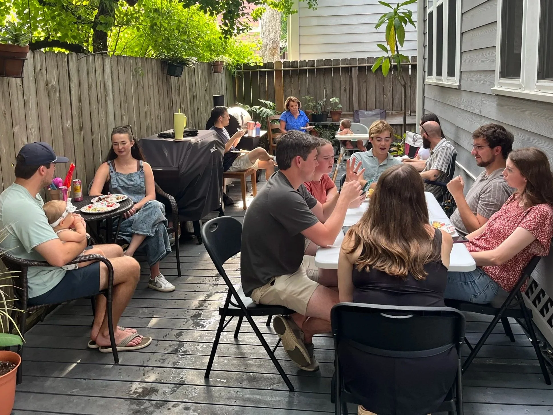 People gathered around a table on a patio, enjoying a meal, with others in the background including children and adults. The setting is outdoors with a wooden fence, house, and greenery.
