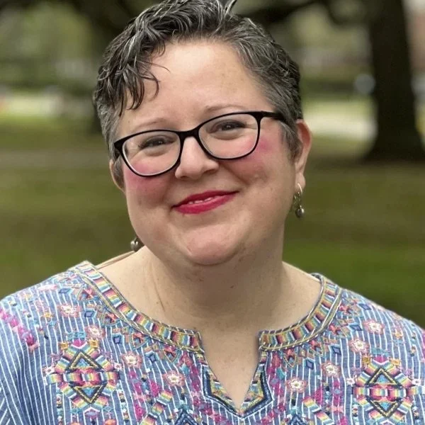 A woman with short curly gray hair, glasses, and earrings smiling outdoors. She is wearing a multicolored embroidered top, and the background shows trees and grass.