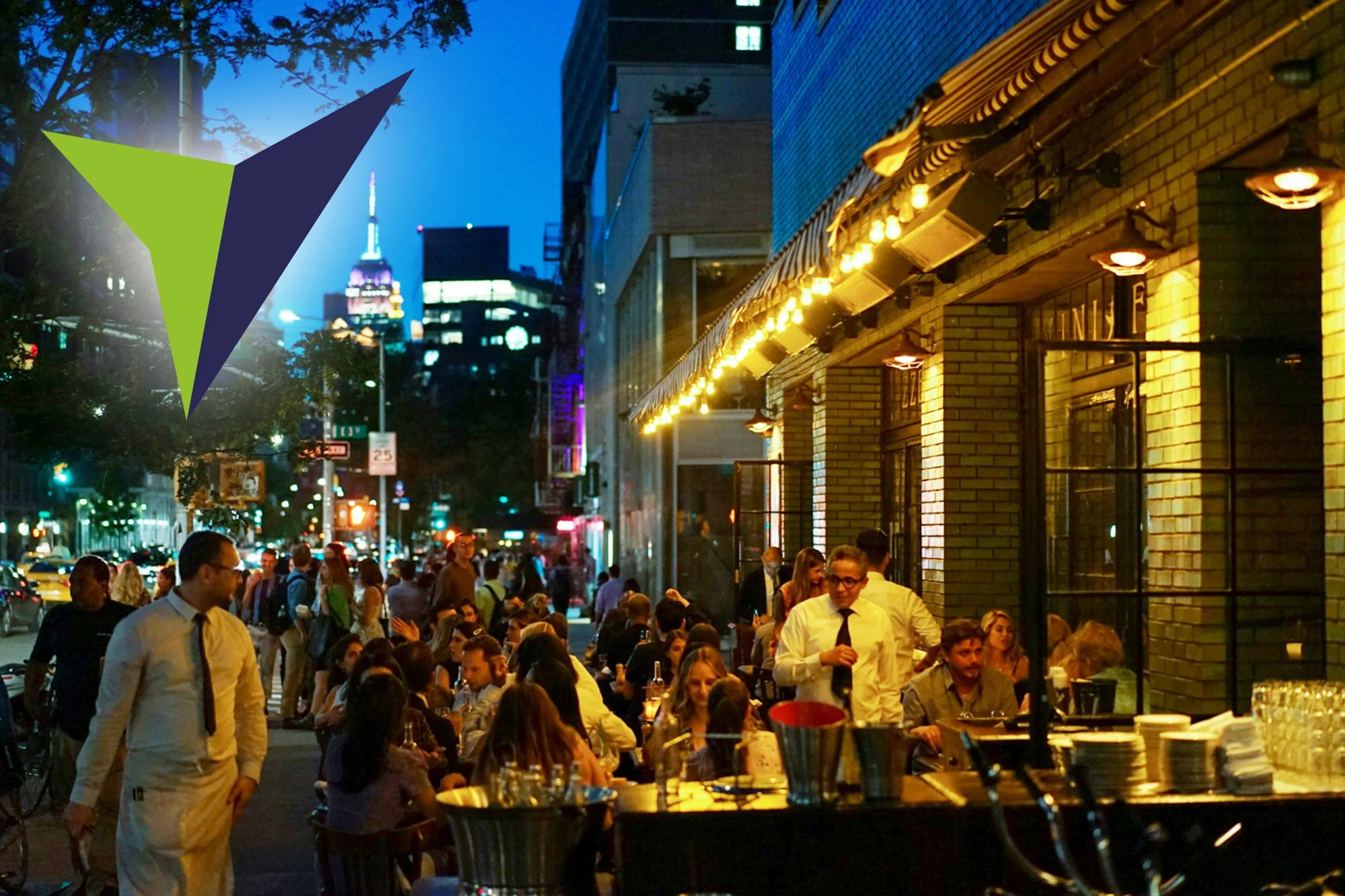 Night scene of a busy city street with a crowded outdoor restaurant, people dining under yellow string lights, and city skyscrapers in the background.