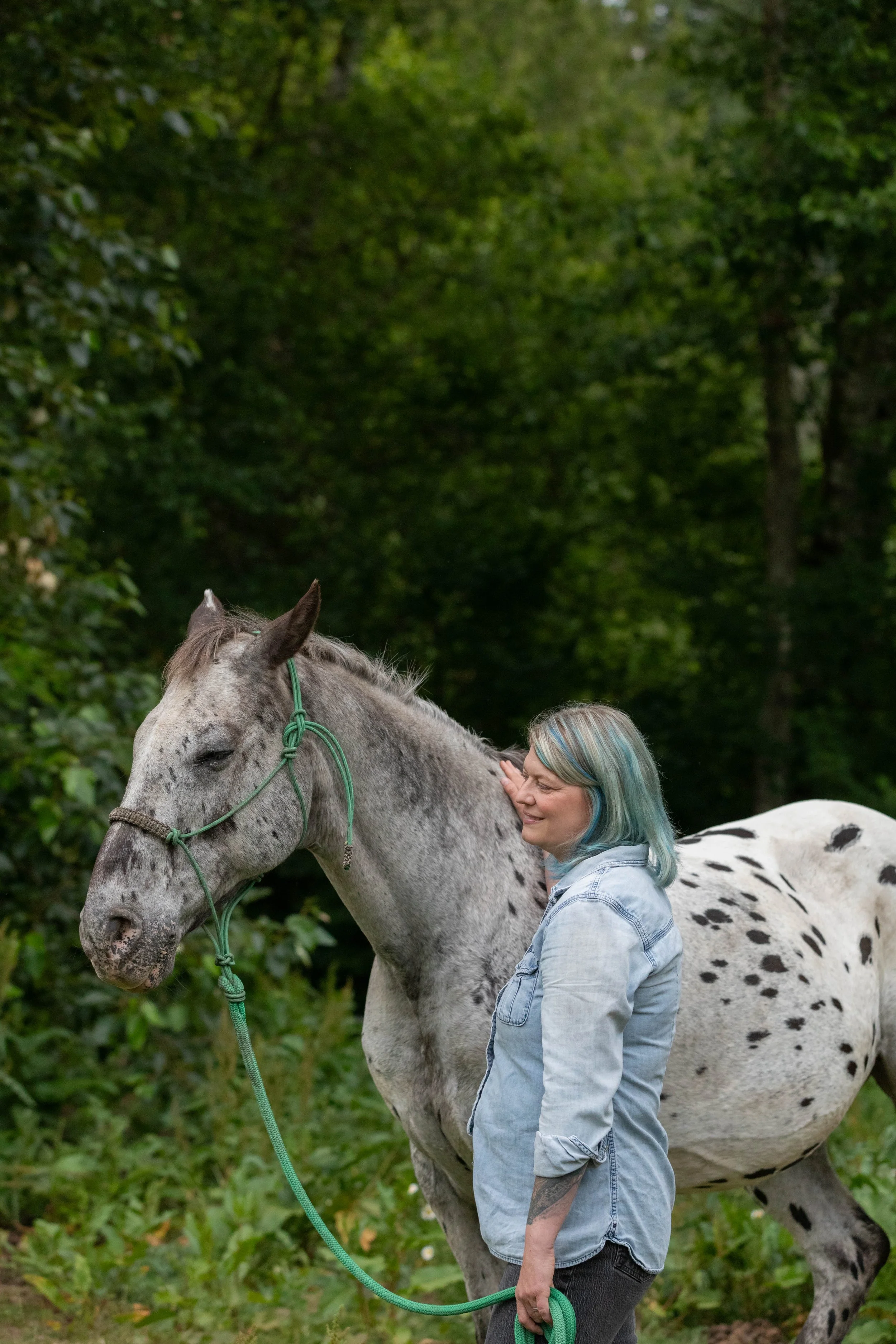 A woman with blue and green hair gently touches the neck of a gray and white-spotted horse in a lush green outdoor setting.