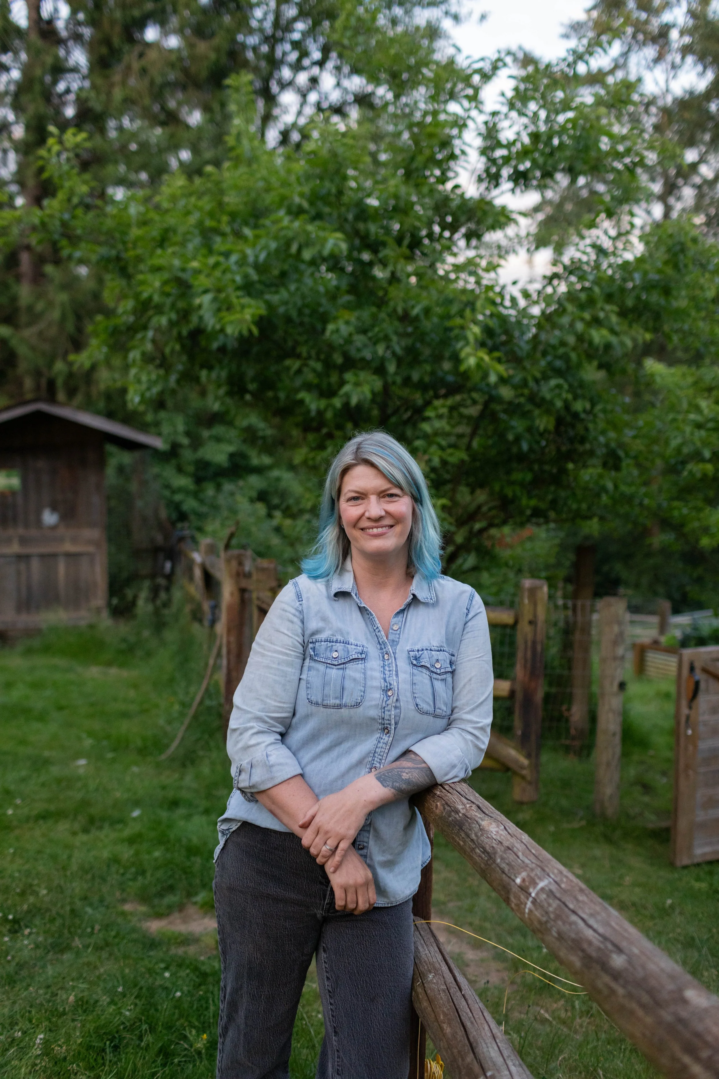 A woman with blue hair standing outdoors, leaning on a wooden fence, with trees and a small wooden shed in the background.