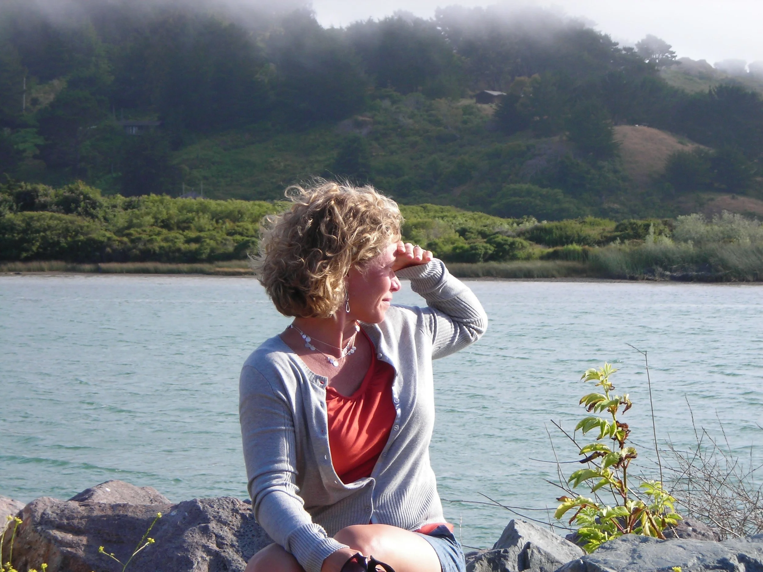 A woman with curly blonde hair sitting on rocks by a body of water in Northern California, shielding her eyes with her hand, with green trees and a hillside in the background.