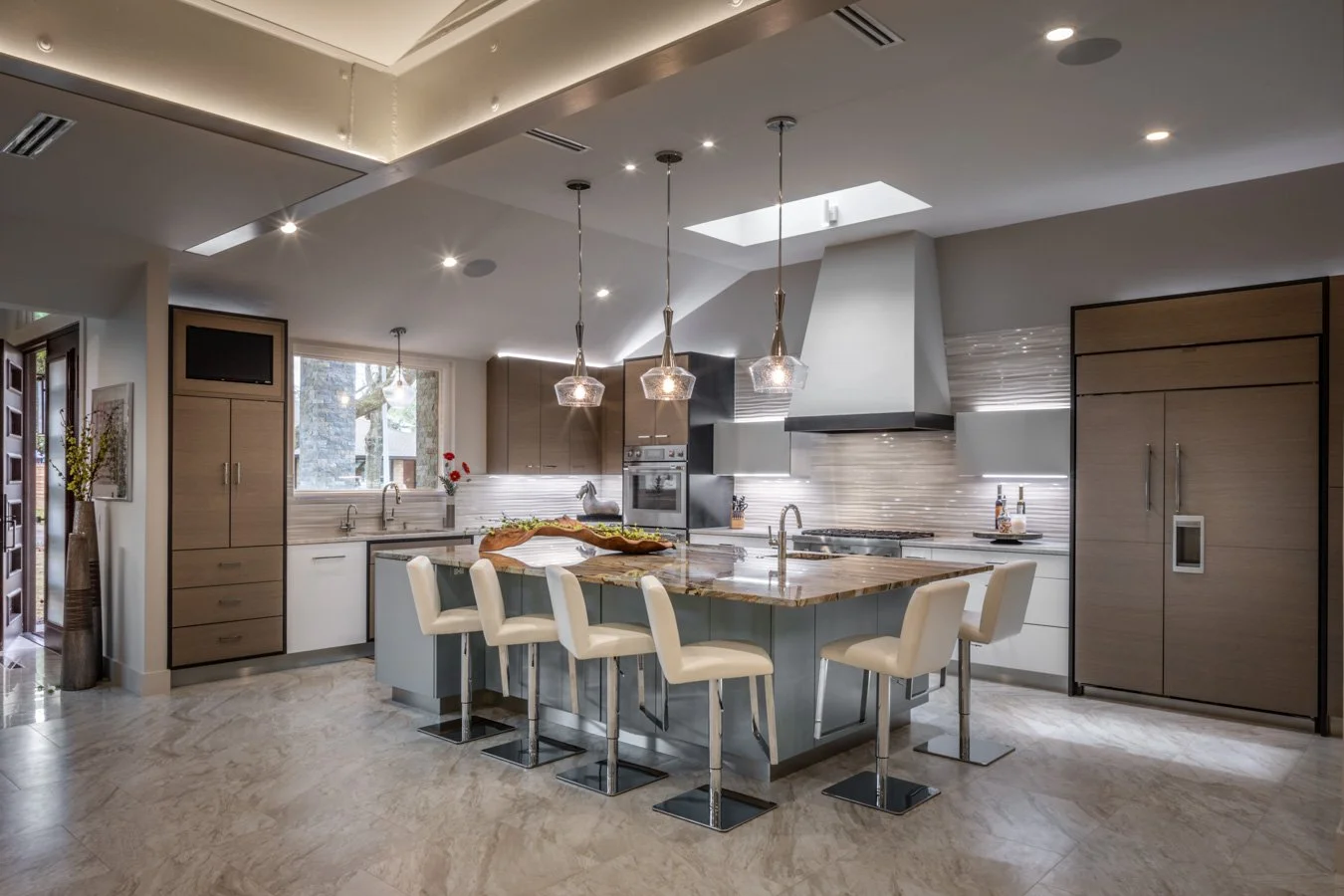 Modern kitchen with beige cabinetry, a marble island with barstools, stainless steel appliances, a window with outdoor view, pendant lights, and a skylight.
