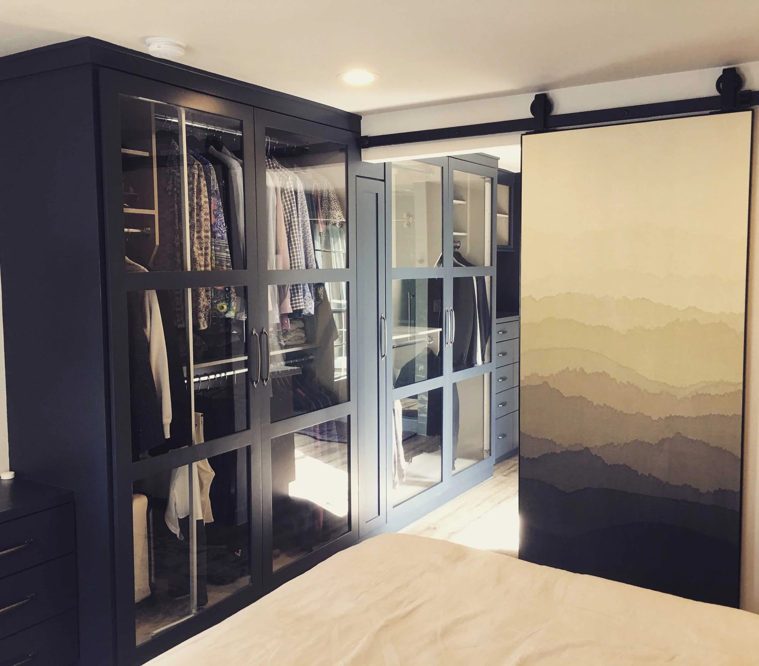 Bedroom with navy blue wardrobe with glass doors, opened to show hanging clothes and shelves, a sliding door to a bathroom, and a decorative folding screen with mountain landscape design.