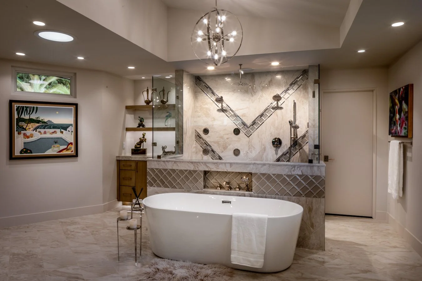 Modern bathroom with a freestanding bathtub, marble walls, decorative shelves with art and sculptures, and a glass-enclosed shower with wall-mounted fixtures, lit by recessed ceiling lights and a circular chandelier.