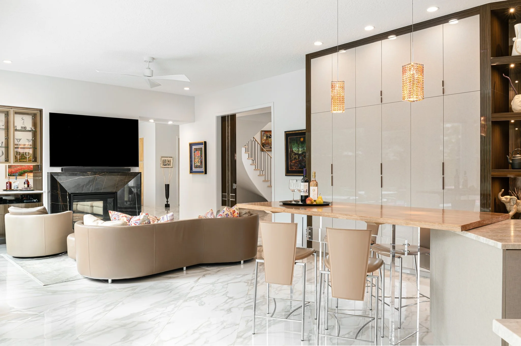 Modern kitchen with white cabinets, a marble tile floor, an island with a wooden countertop and pendant lights looking towards the family TV area and custom marble fireplace.