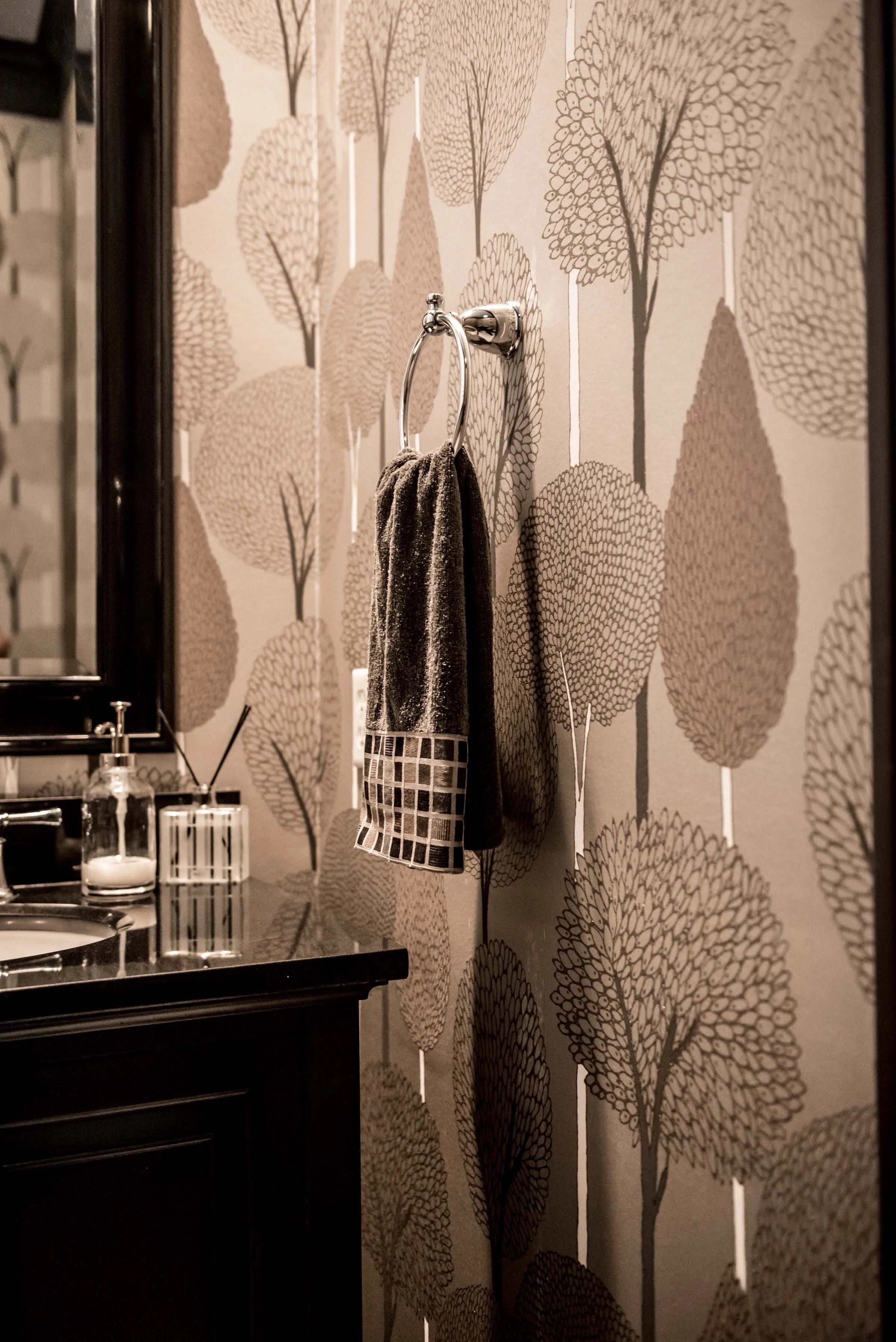 A bathroom with wallpaper featuring a pattern of large, stylized trees in shades of brown and beige. There is a dark wood vanity with a black countertop, a small glass bottle, and a candle. A silver towel ring with a dark towel and a checkered cloth hangs on the wall.