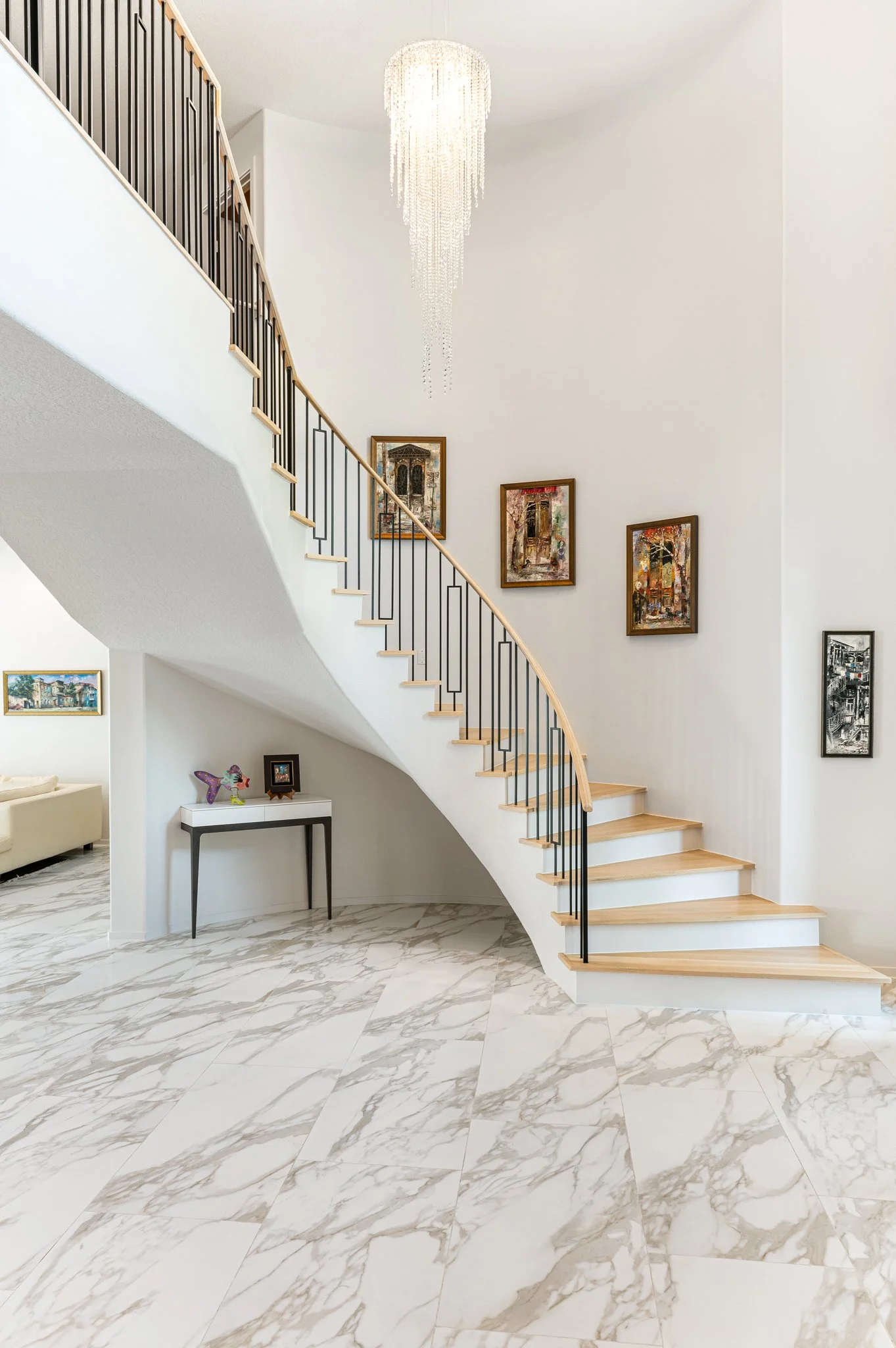 A modern interior featuring a curved staircase with wooden steps and black metal rails, marble flooring, a white wall with colorful framed artwork, a chandelier hanging from the ceiling, and a small table with decorative items in the corner.