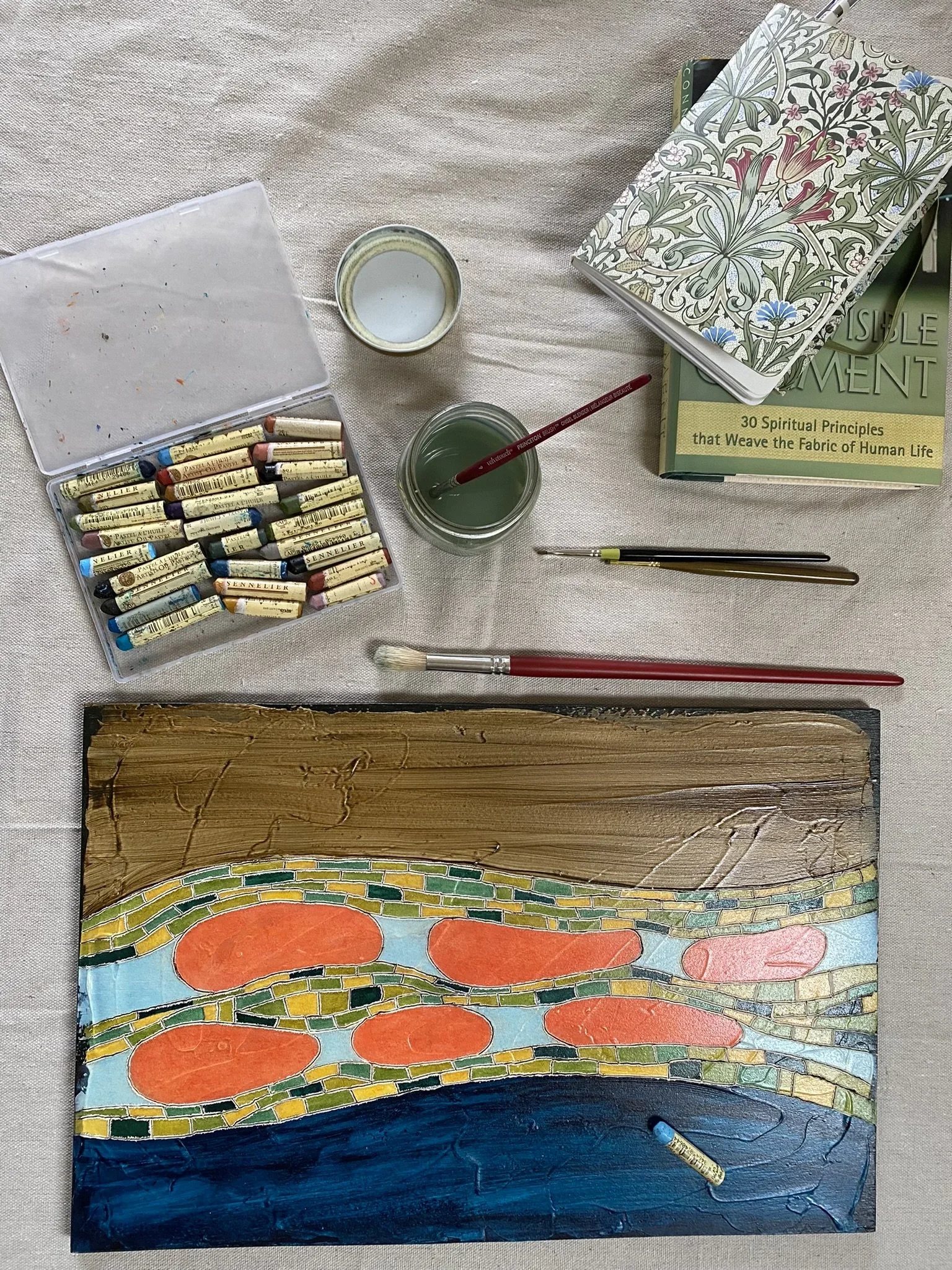A colorful painting of a mosaic design featuring large orange circles, smaller green and yellow shapes, and blue waves at the top. The painting is on a wooden board, resting on a surface with art supplies nearby, including a glue stick and paintbrush.