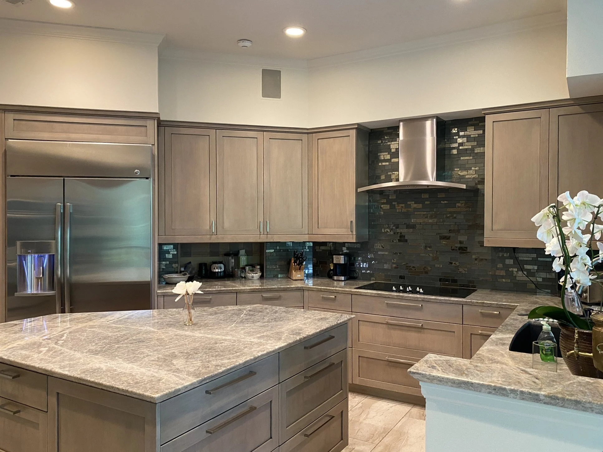 Modern kitchen with grey washed Woodmode cabinets, granite island, stainless steel appliances, a black glass backsplash and a white orchid.