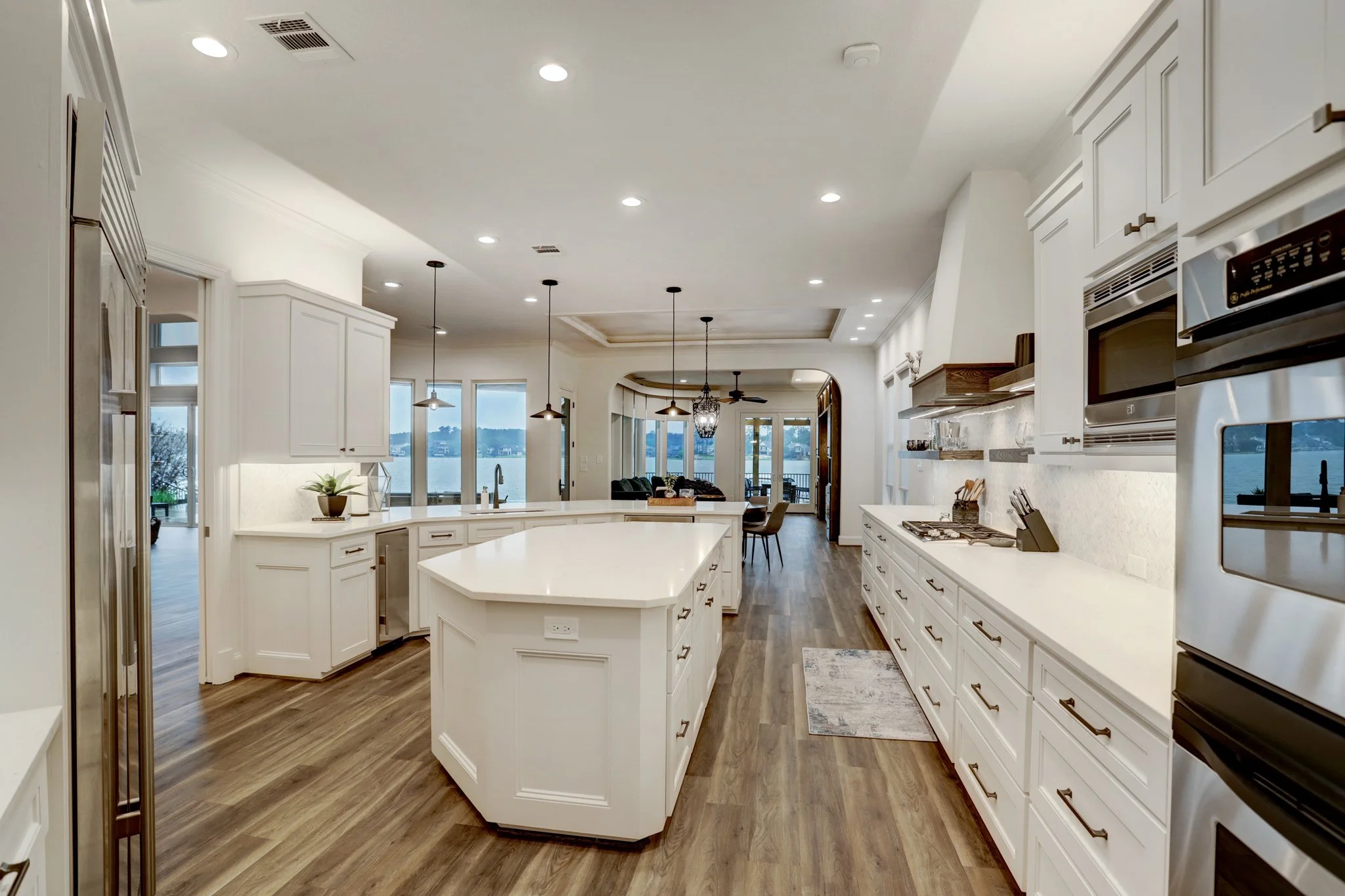 Modern kitchen with white cabinets, wood flooring, and large windows overlooking a body of water.