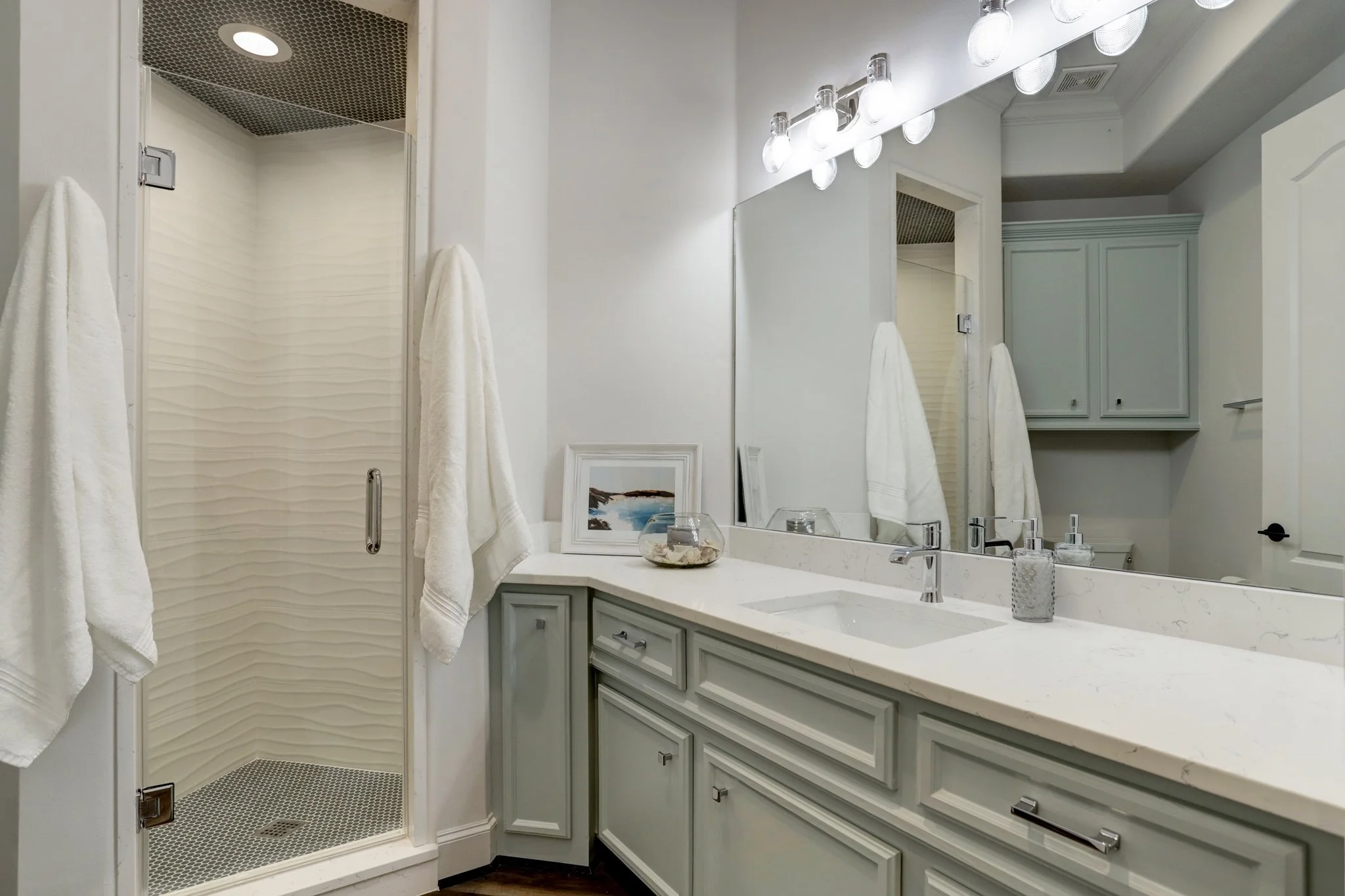 A clean bathroom with a light-colored vanity, a large mirror, bright overhead lights, towels hanging, a framed picture, and a shower stall with textured walls.