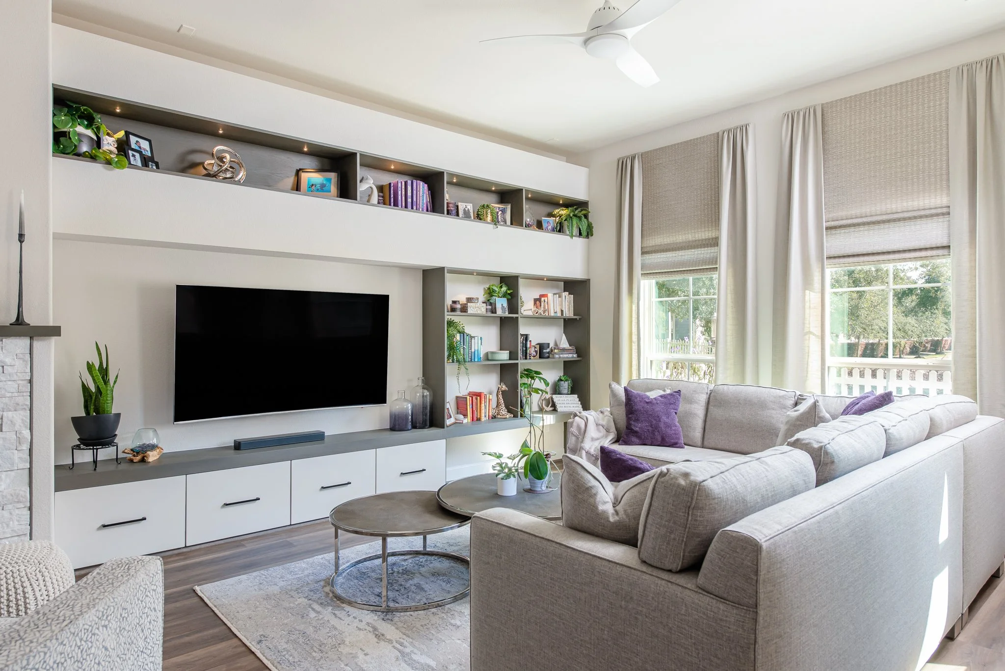 Living room with a beige sectional sofa with purple pillows, a television mounted on a gray and white built-in unit, and large windows with beige curtains.