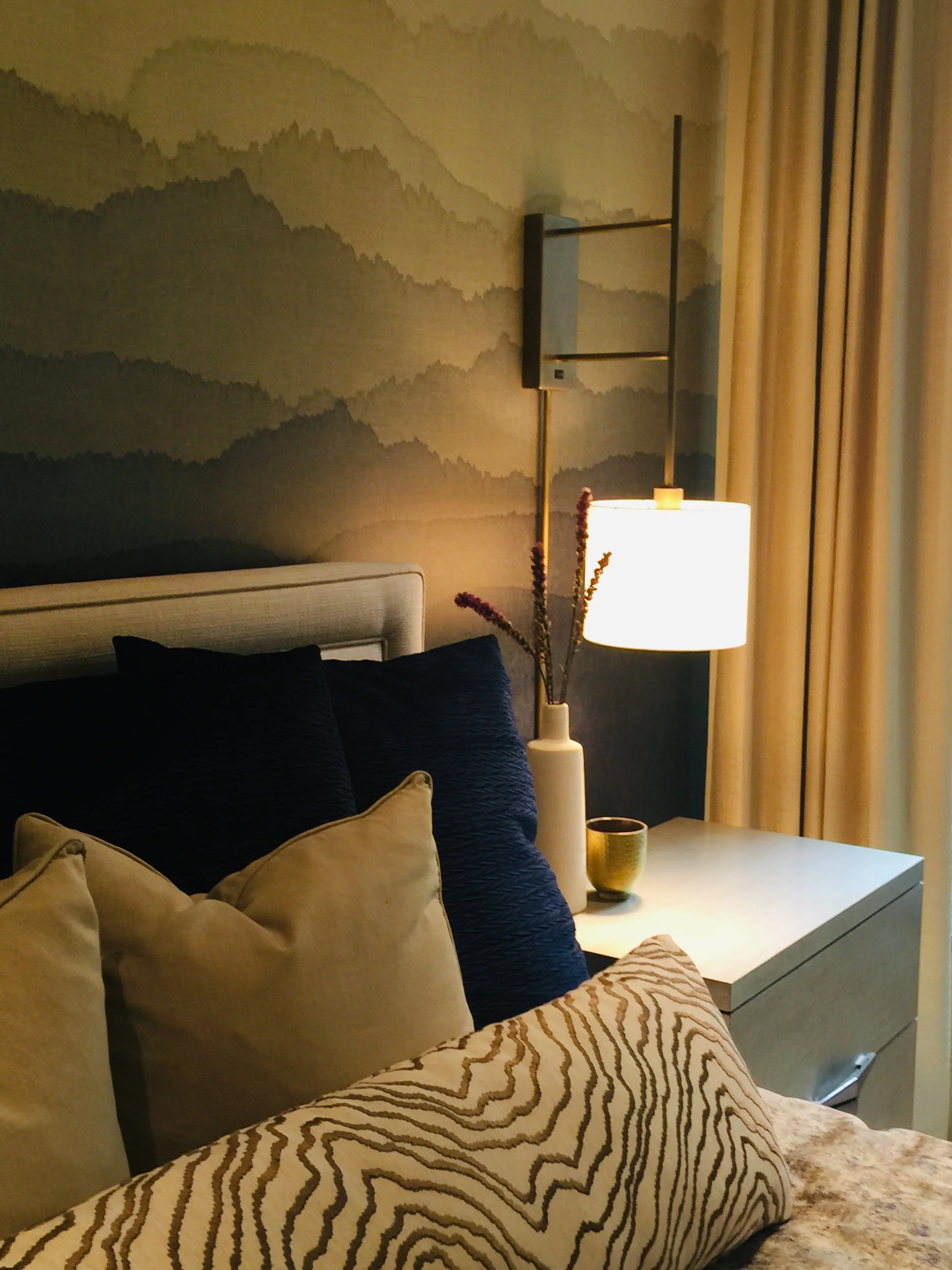 A bedroom corner with a nightstand, a lamppost, and a bed with beige and dark blue pillows. The wall features a mountain landscape mural. There are beige curtains to the side.