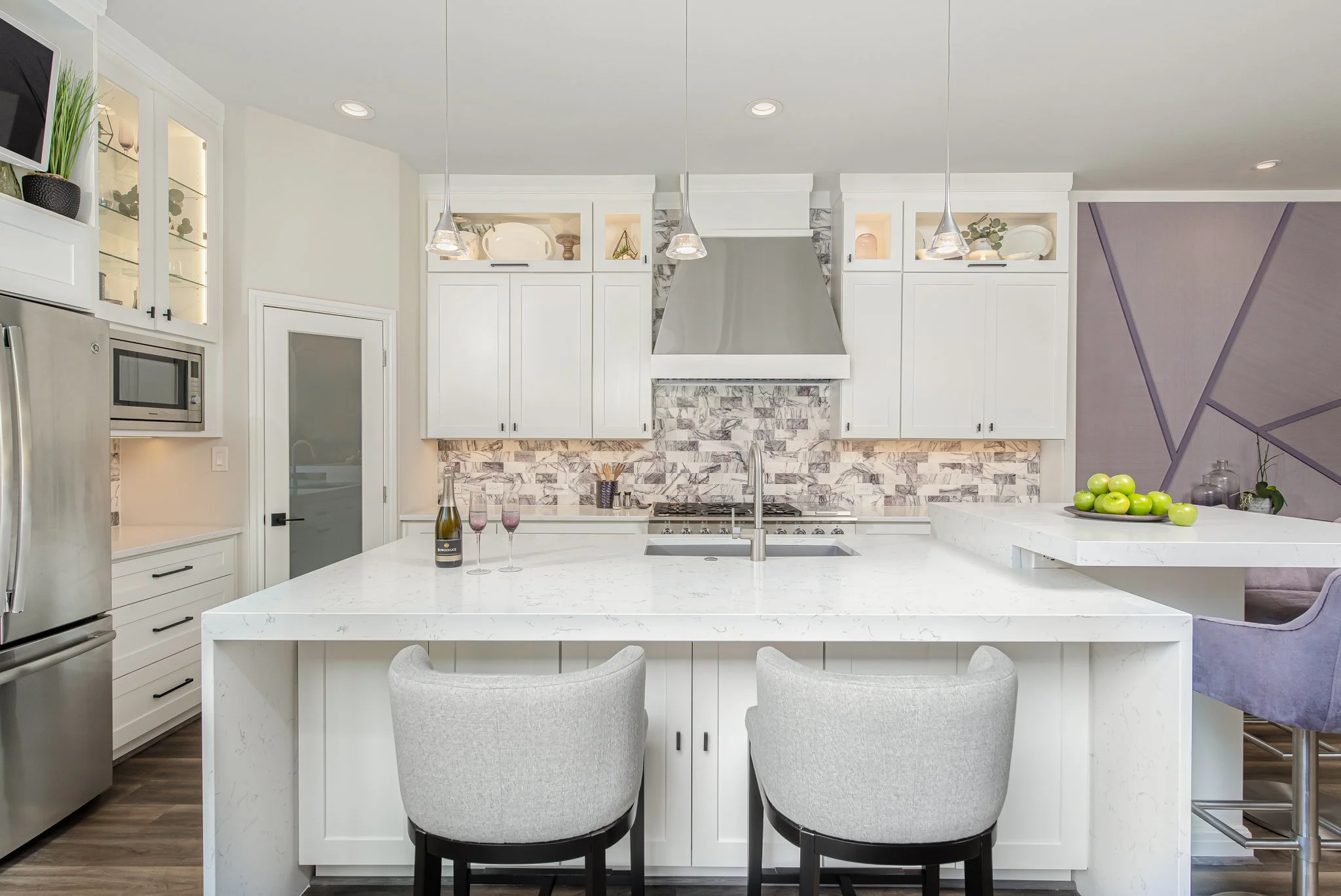 Modern kitchen with white cabinets, marble island, stainless steel appliances, a backsplash with gray and white mosaic tiles, and a purple accent wall, decorated with plants, glassware, and a bowl of green apples.
