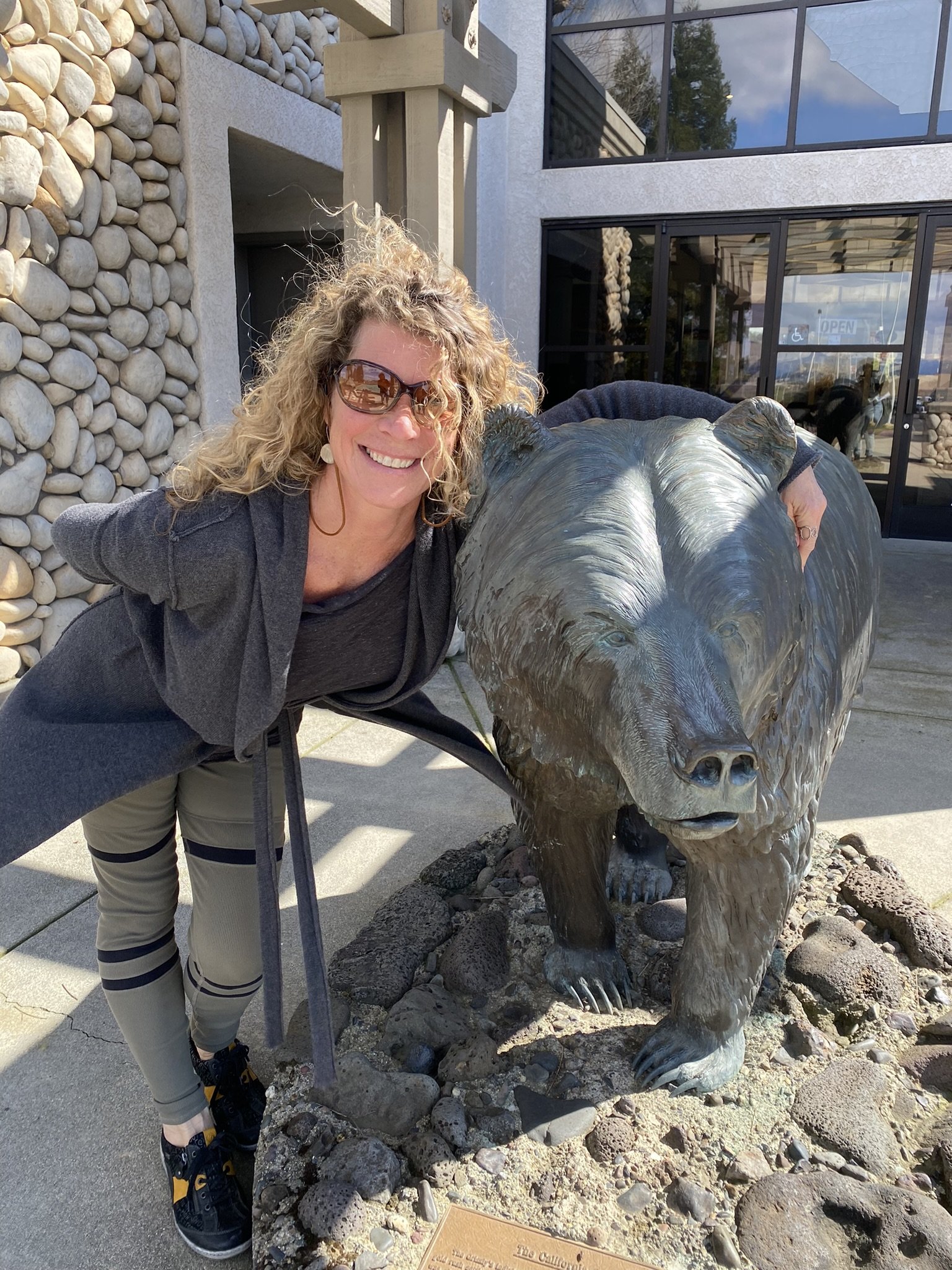 Woman smiling with sunglasses leaning on a bear sculpture outside a building with a pebble and rock landscaping.