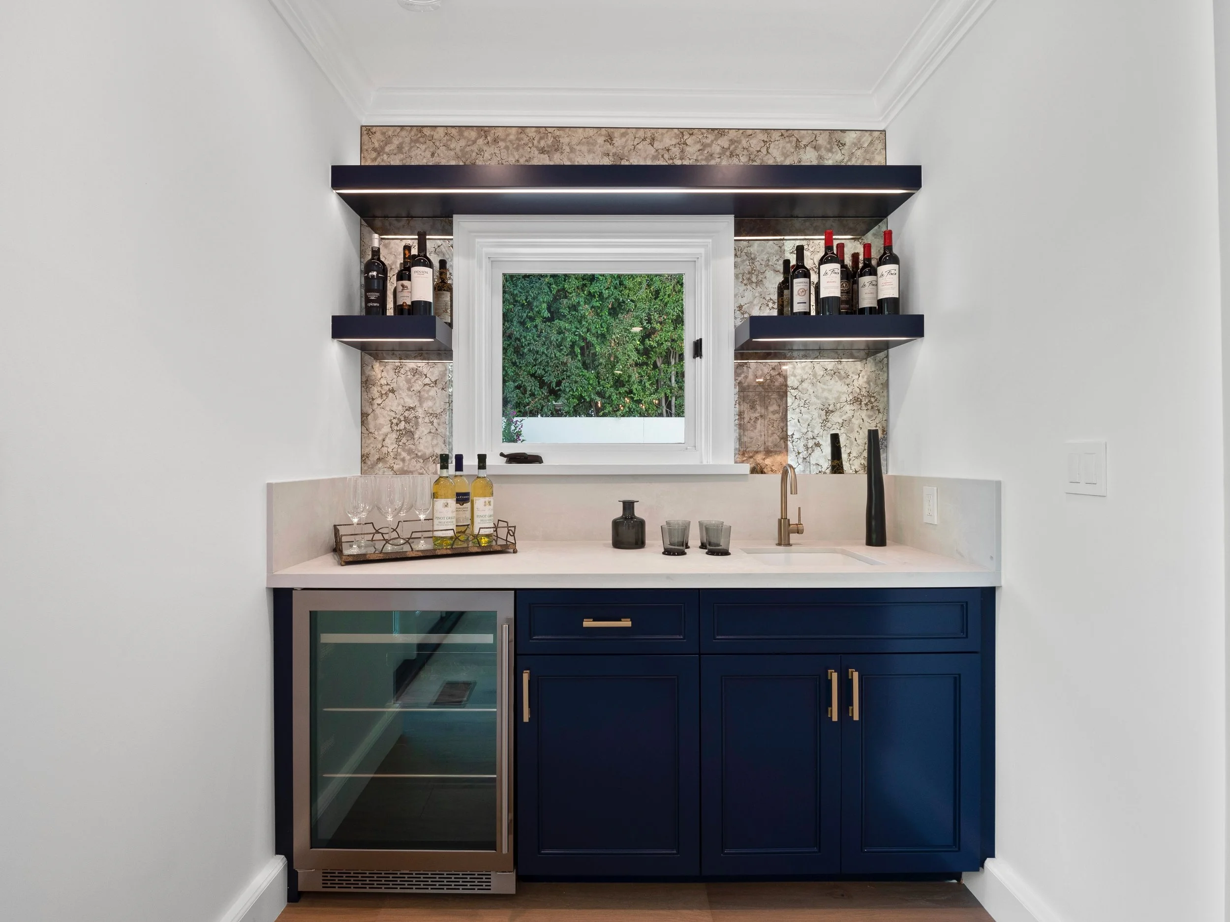 Hancock Park custom bar with navy cabinetry, quartz countertop, undercounter wine fridge, brass faucet, and backlit stone backsplash with floating shelves.