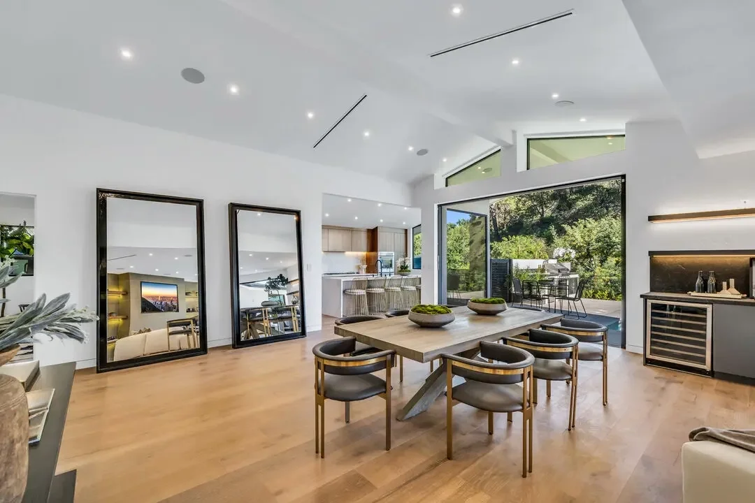 Spacious modern dining area with a wooden table, eight black and gold chairs, large mirrors, and sliding glass doors leading to a balcony with outdoor seating and green trees.