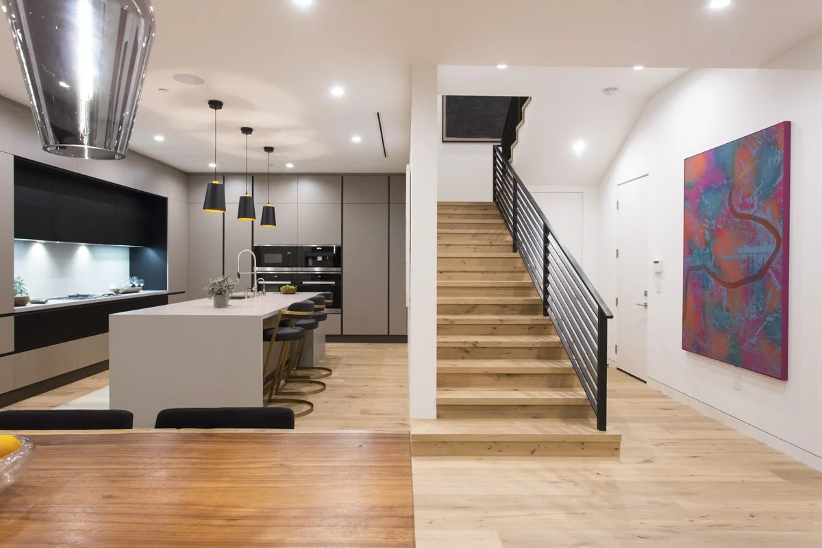 Open-concept kitchen in Venice modern home with matte taupe cabinetry, integrated appliances, waterfall island, and floating oak staircase with black steel railing.