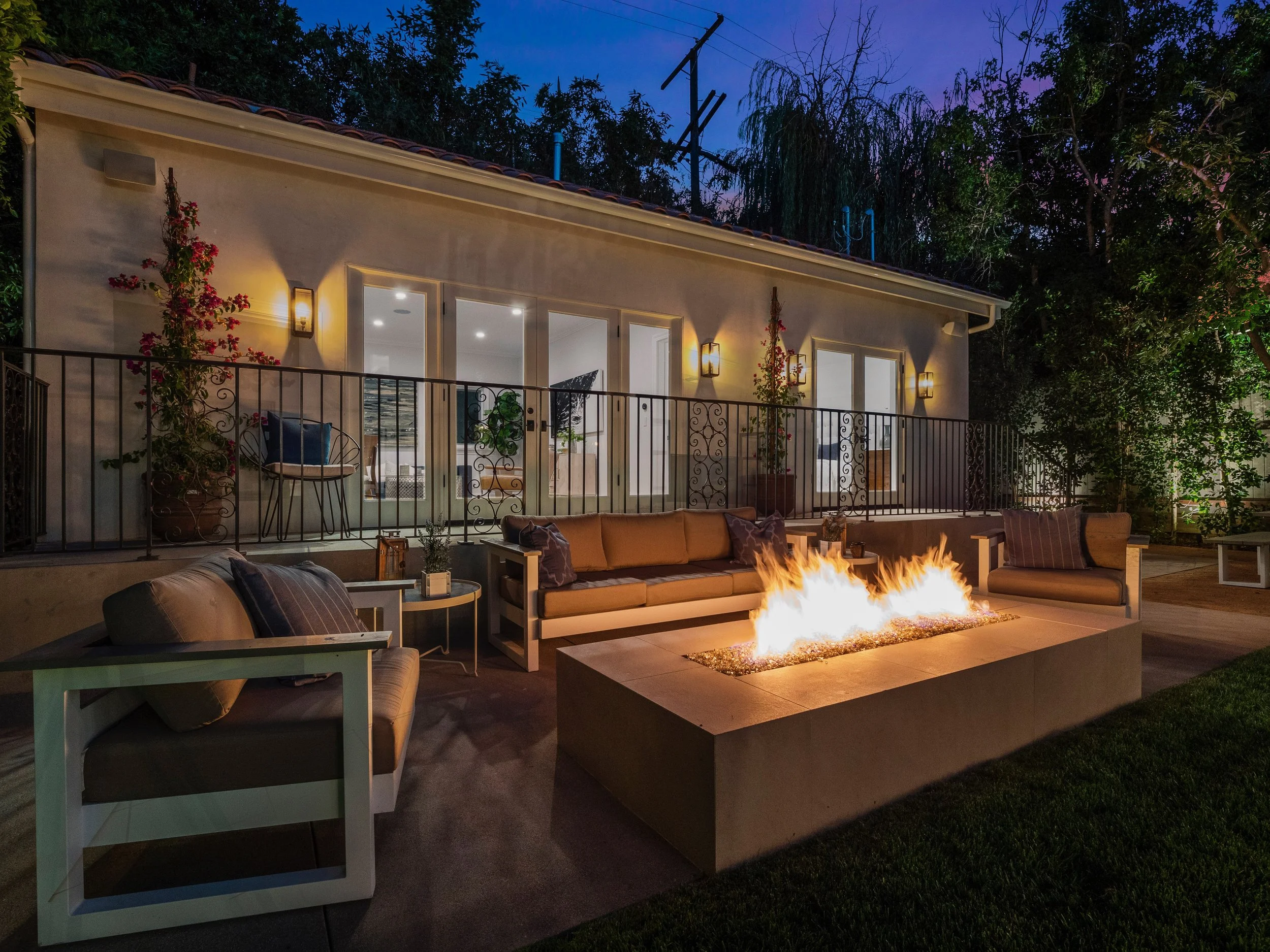 Hancock Park backyard at dusk with modern fire pit lounge, outdoor seating, wrought iron balcony, and softly lit Spanish-style home exterior.