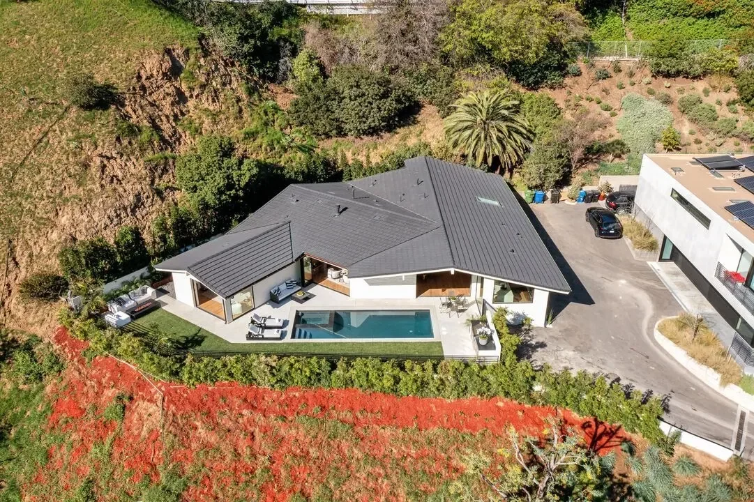 Aerial view of a modern house with a swimming pool, outdoor seating areas, and a parking lot surrounded by greenery and a hillside.