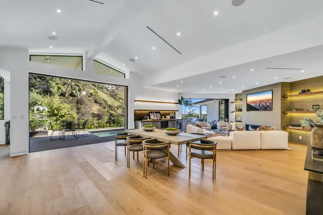 Modern living room with large windows, open floor plan, white sofa, wooden dining table with chairs, built-in shelves, and scenic outdoor view.