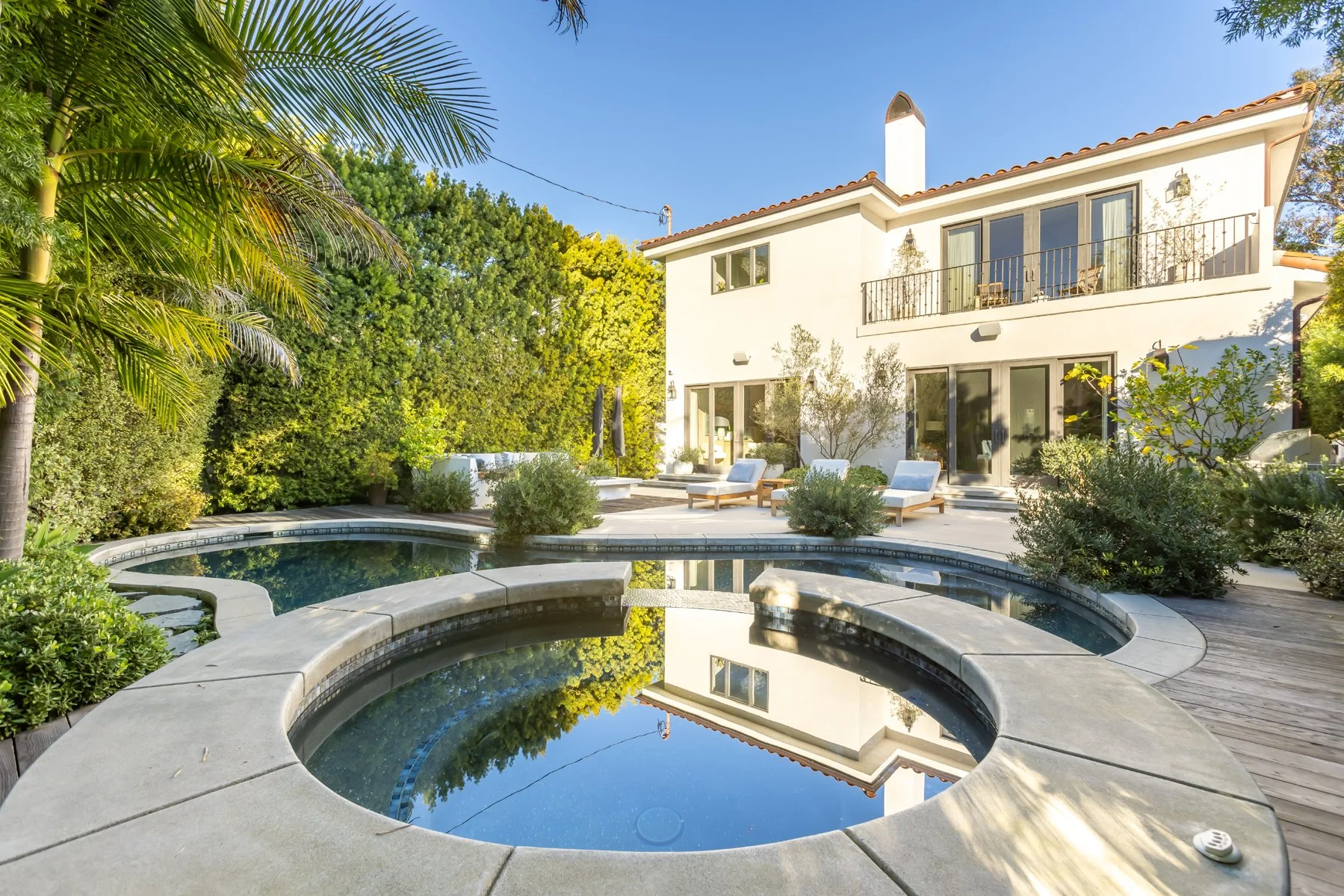 Backyard of a custom Brentwood home featuring a curved swimming pool, wood deck, and Spanish-style residence surrounded by lush landscaping.