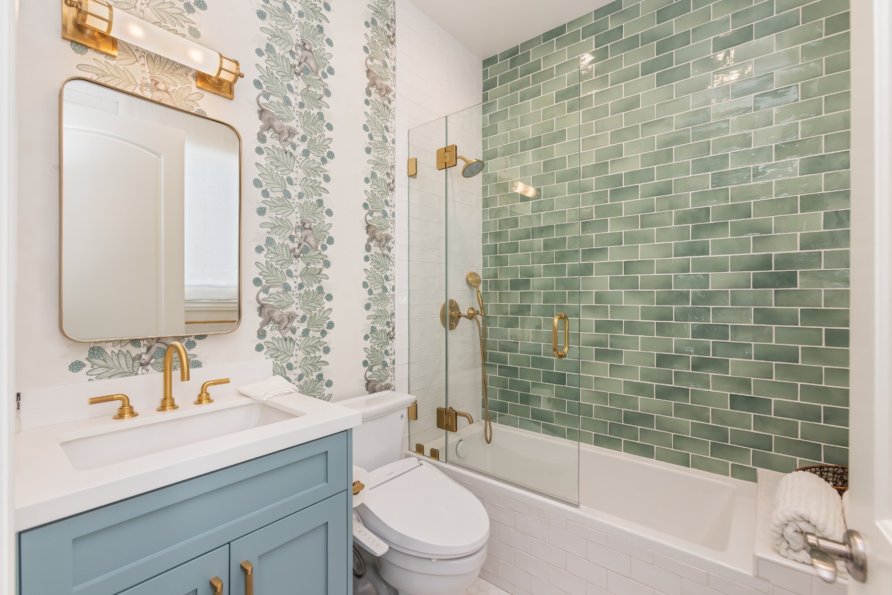 Guest bathroom in a custom Brentwood home featuring green subway tile tub surround, patterned wallpaper, blue vanity, brass fixtures, and a glass shower enclosure.