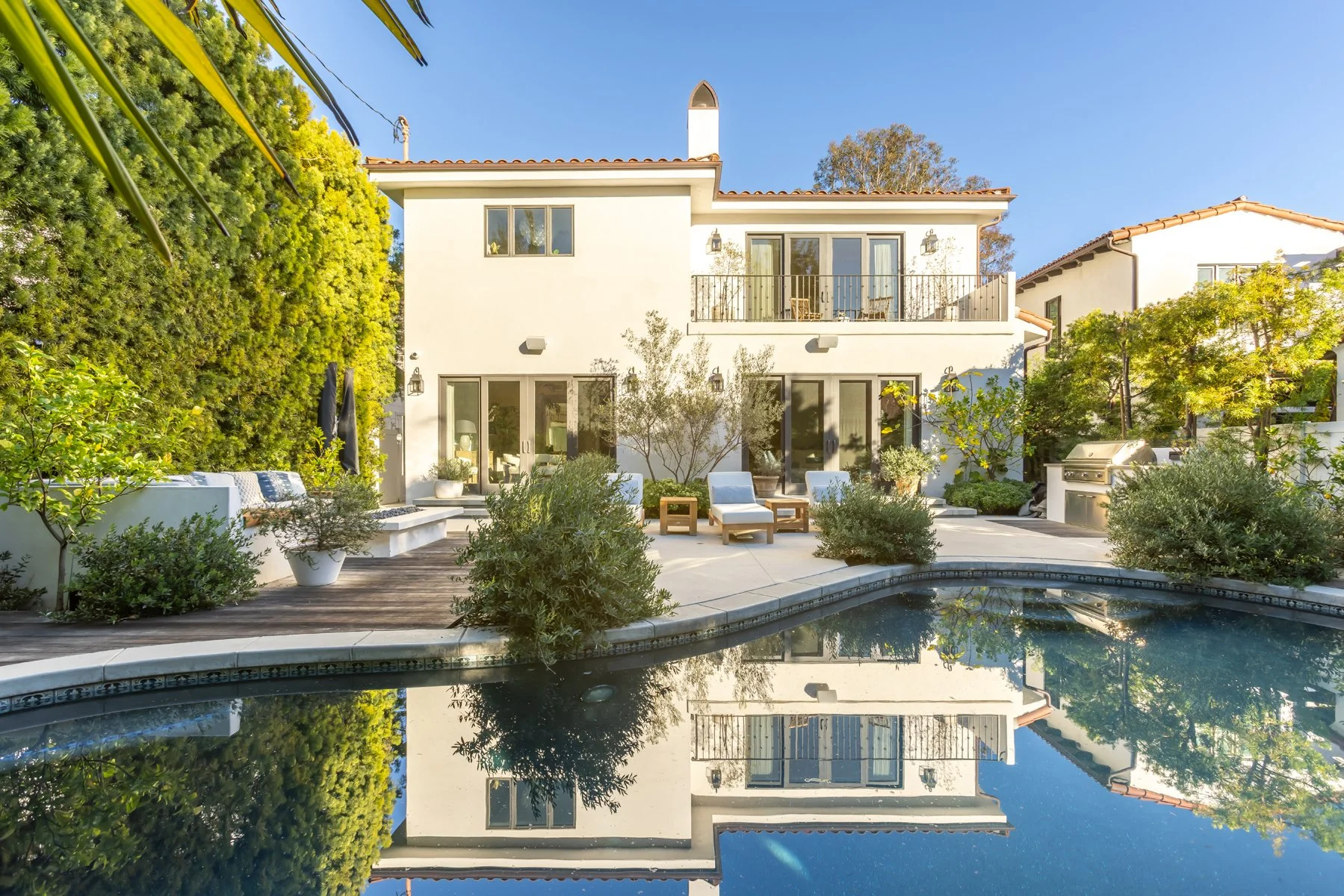 Backyard of a custom Brentwood home featuring a Spanish-style two-story residence, swimming pool with reflective water, outdoor lounge seating, mature landscaping, and a second-floor balcony.
