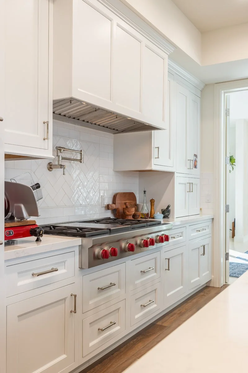 Kitchen range wall in a custom Brentwood home featuring white shaker cabinetry, a professional gas range with red control knobs, herringbone tile backsplash, integrated vent hood, and stone countertops.
