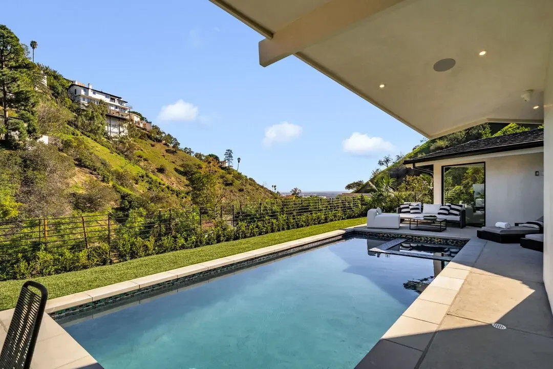 Modern outdoor patio with a swimming pool, lounge furniture, and scenic hillside view under a clear sky.