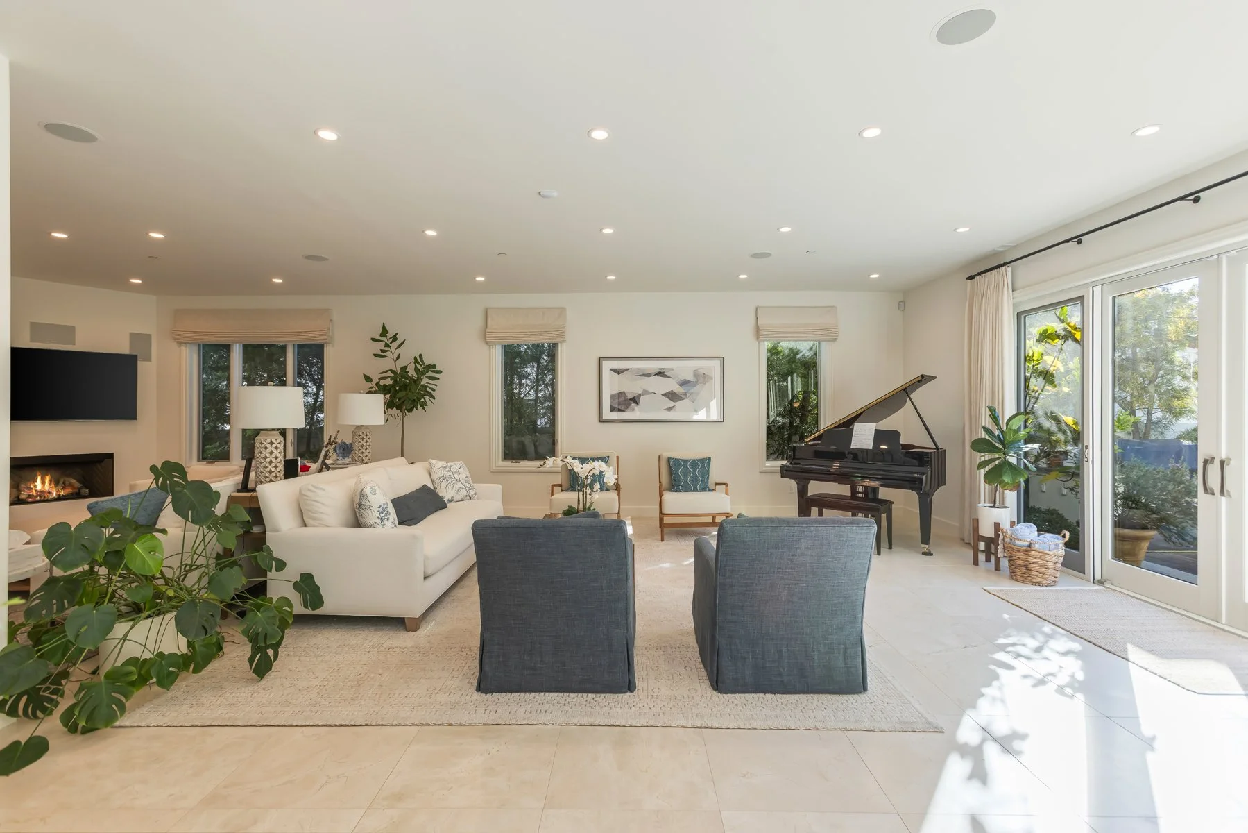 Spacious living room in a custom Brentwood home featuring a fireplace, grand piano, light stone flooring, recessed ceiling lights, neutral furnishings, and large glass doors opening to the garden.