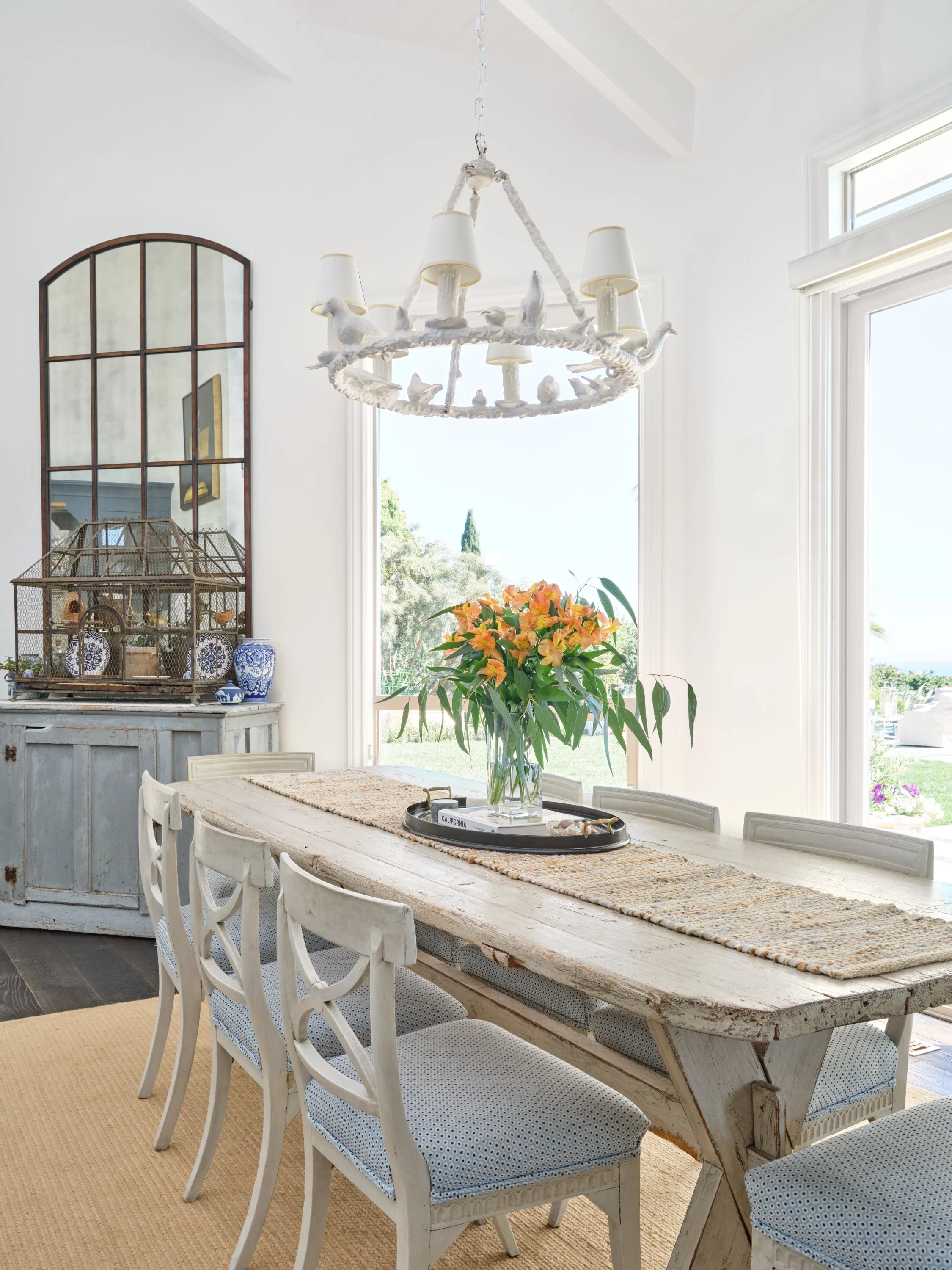 Malibu custom home dining room with rustic wood table, white chairs, statement chandelier, and large windows overlooking the garden by Heart Construction