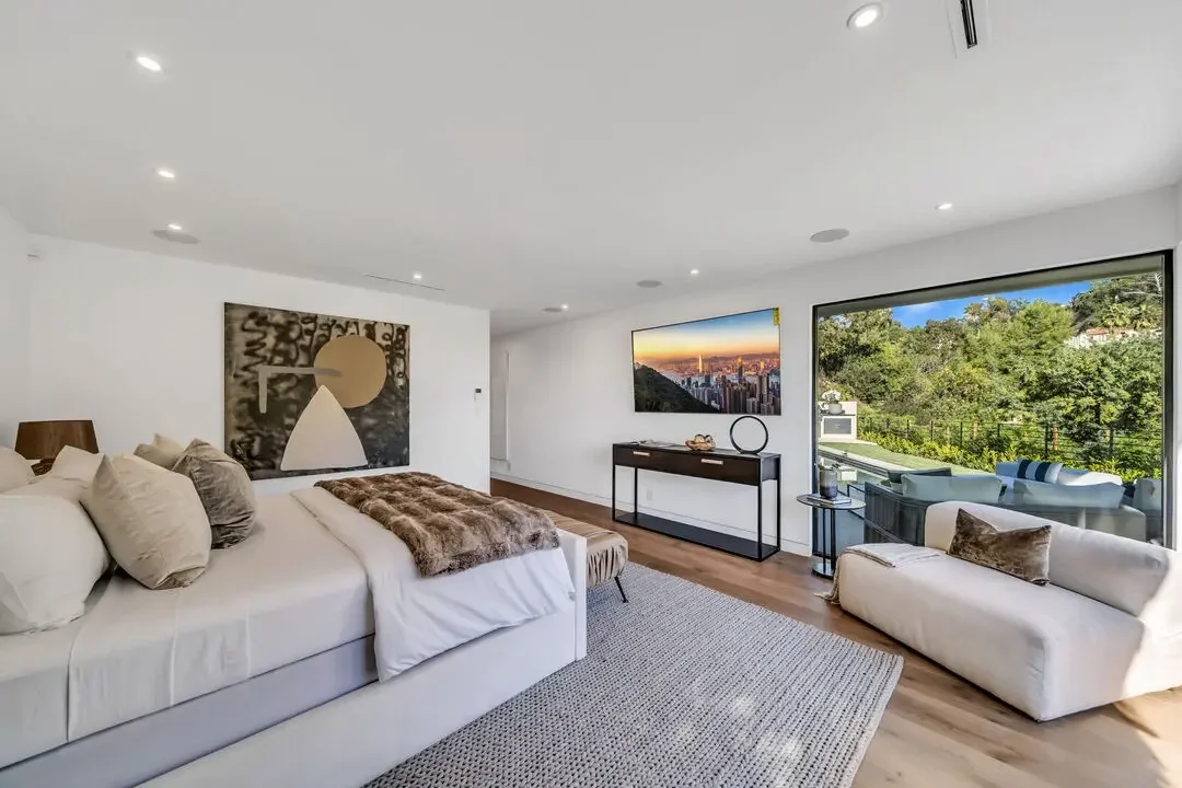 Modern bedroom with large bed, white and beige furniture, artwork on the wall, and a view of a green outdoor area through sliding glass doors.