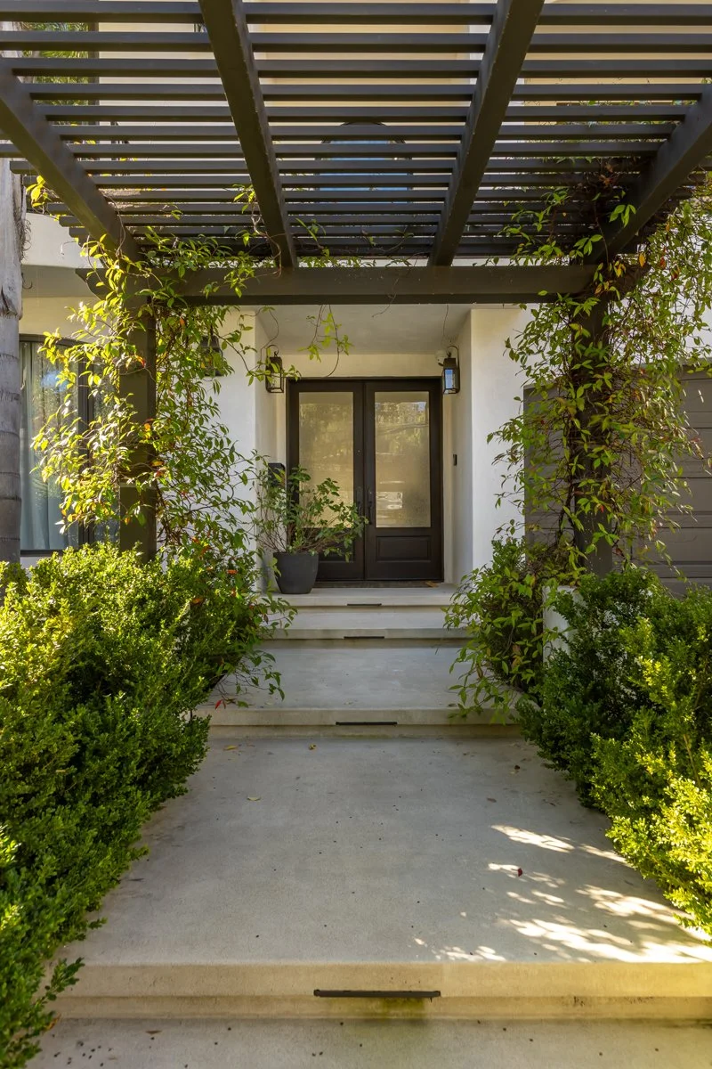 Front entry of a custom home in Brentwood, Los Angeles, featuring a modern wood pergola, black-framed glass entry door, concrete steps, and lush climbing greenery.