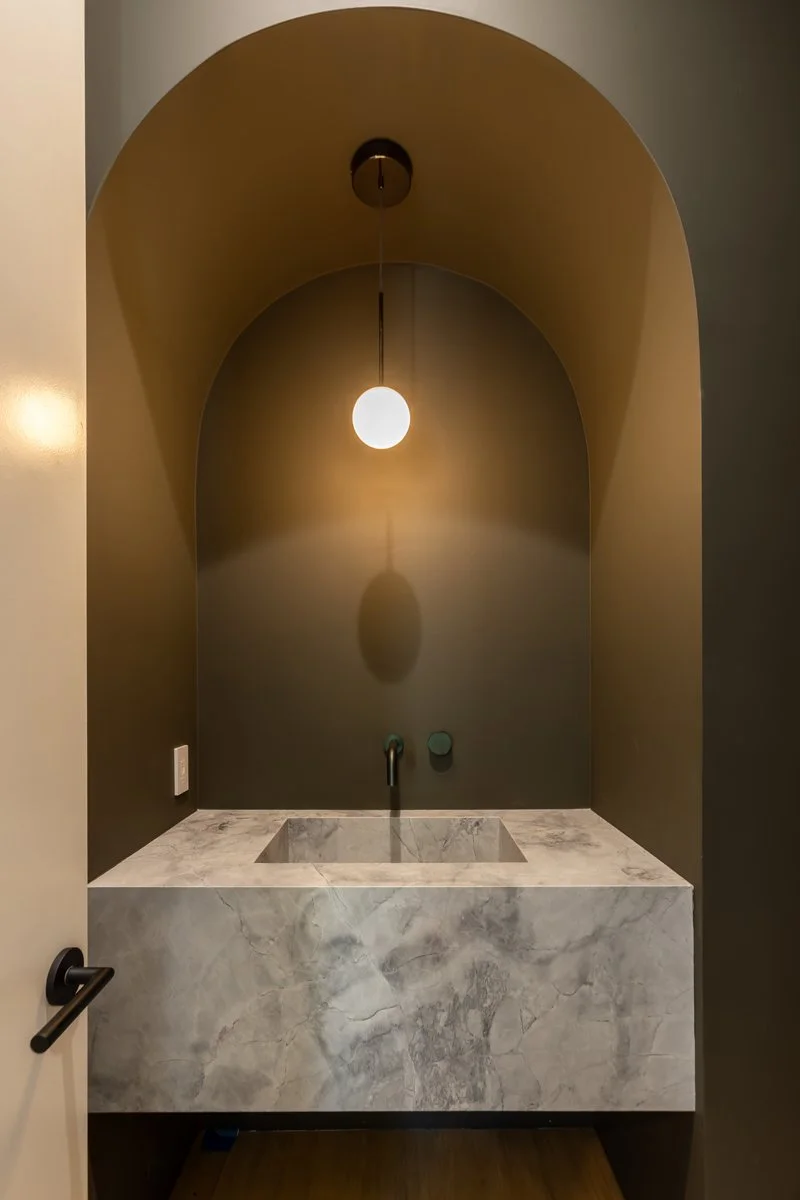 Custom arched powder room vanity with integrated marble sink, warm accent lighting, and minimalist brass pendant in a modern Mar Vista home remodel by Heart Construction.