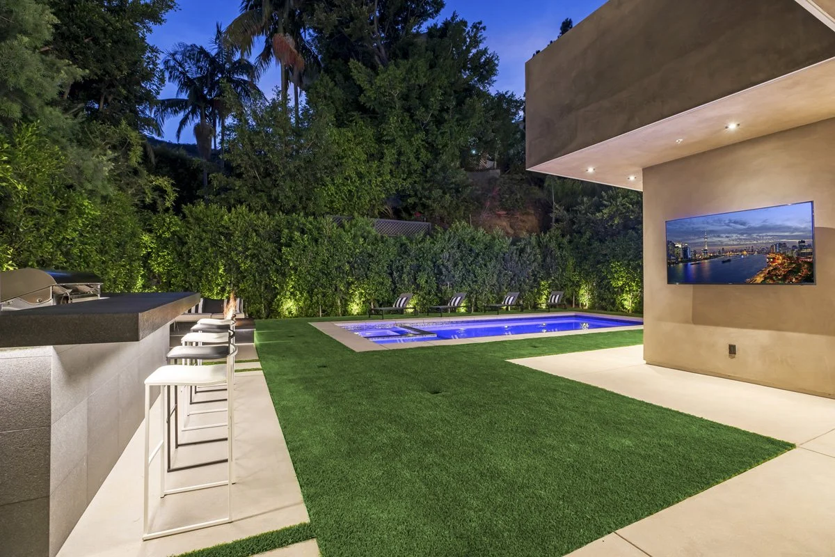 Beverly Hills outdoor kitchen and pool at dusk with bar seating, artificial turf lawn, and perimeter hedge lighting