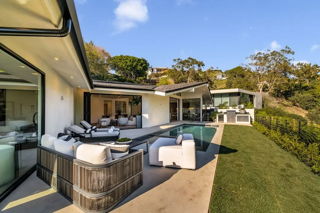 Modern house with a backyard pool, outdoor seating, and a grassy yard, surrounded by trees and hills under a blue sky.