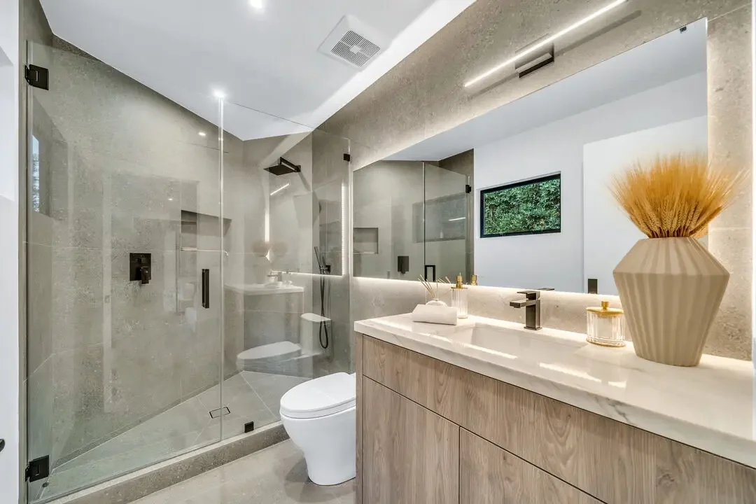 Modern bathroom with a glass-enclosed shower, a white toilet, and a wooden vanity with a marble countertop. There is a large mirror above the sink, a small window showing greenery, and decorative items including a vase with dried plants.