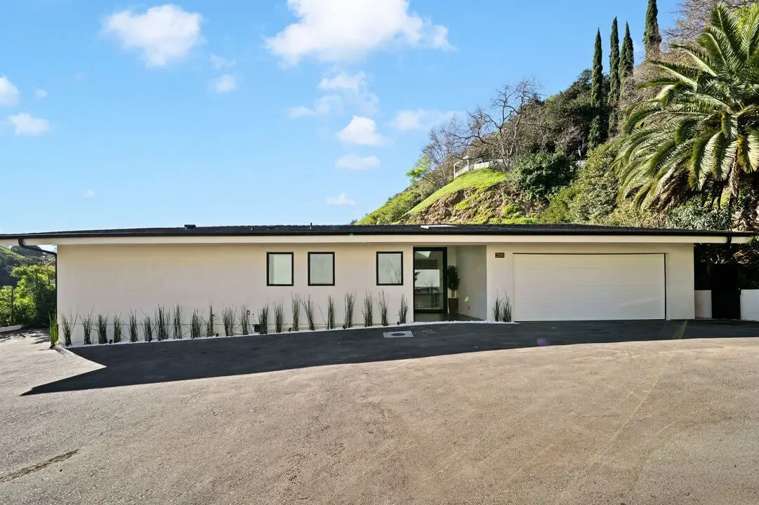 Modern single-story house with white exterior, black window frames, a large garage door, and landscaped front yard with tall grass plants. Hills with trees are visible in the background under a partly cloudy sky.