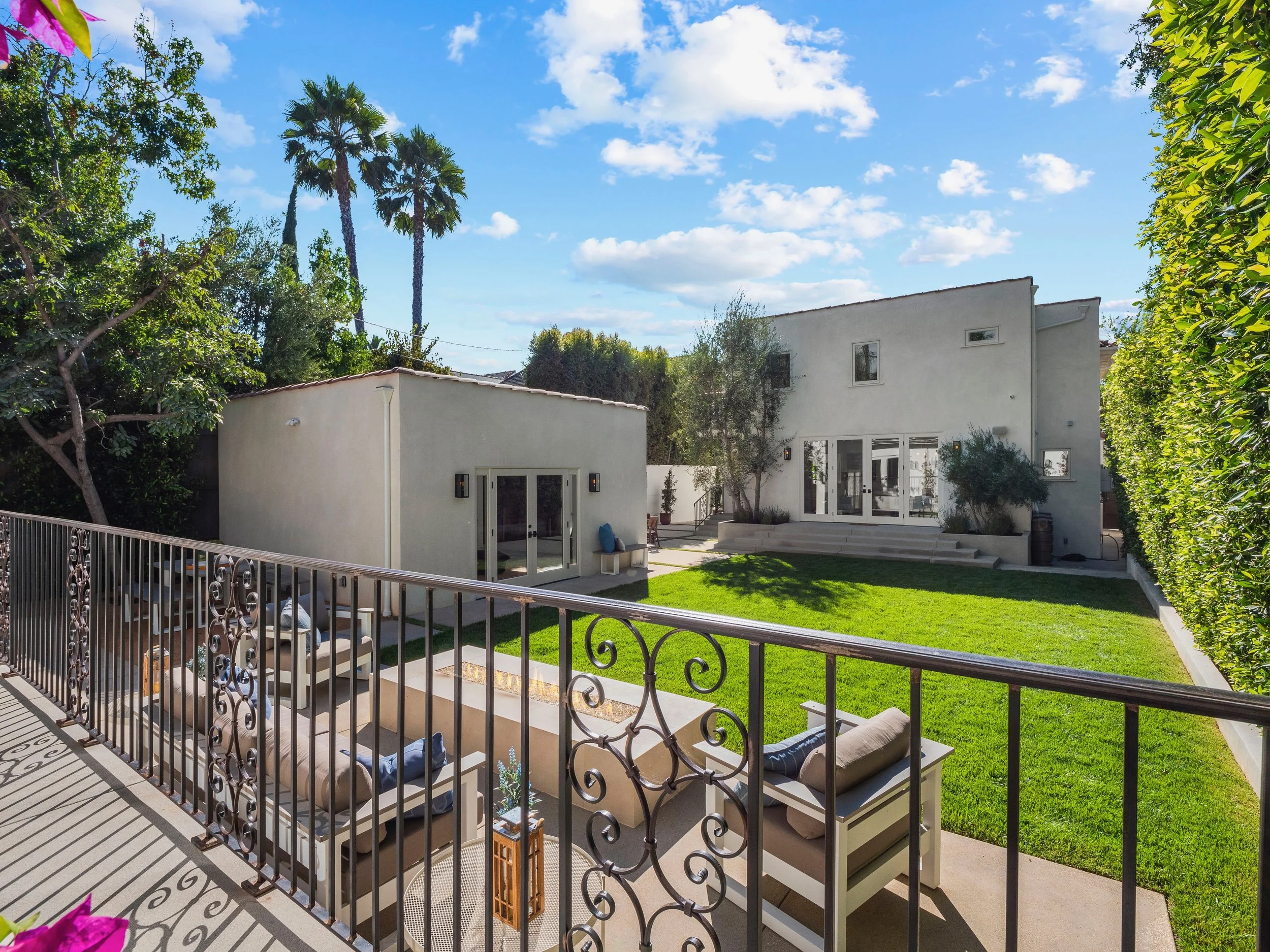 Hancock Park backyard with detached guest house, lush lawn, outdoor fire pit lounge, and wrought iron balcony overlooking the Spanish-style residence.