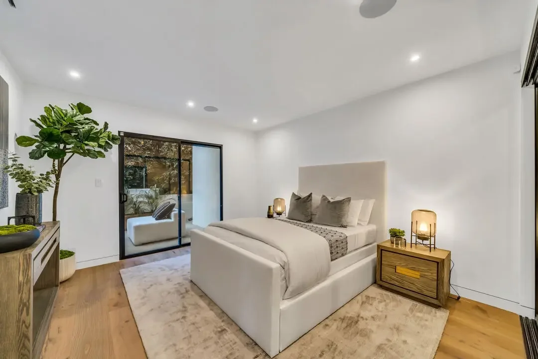 Modern bedroom with white walls, a large bed with beige or light gray bedding, wooden nightstand with a lamp and small plants, large sliding glass door leading to a balcony, and a potted plant next to the door.